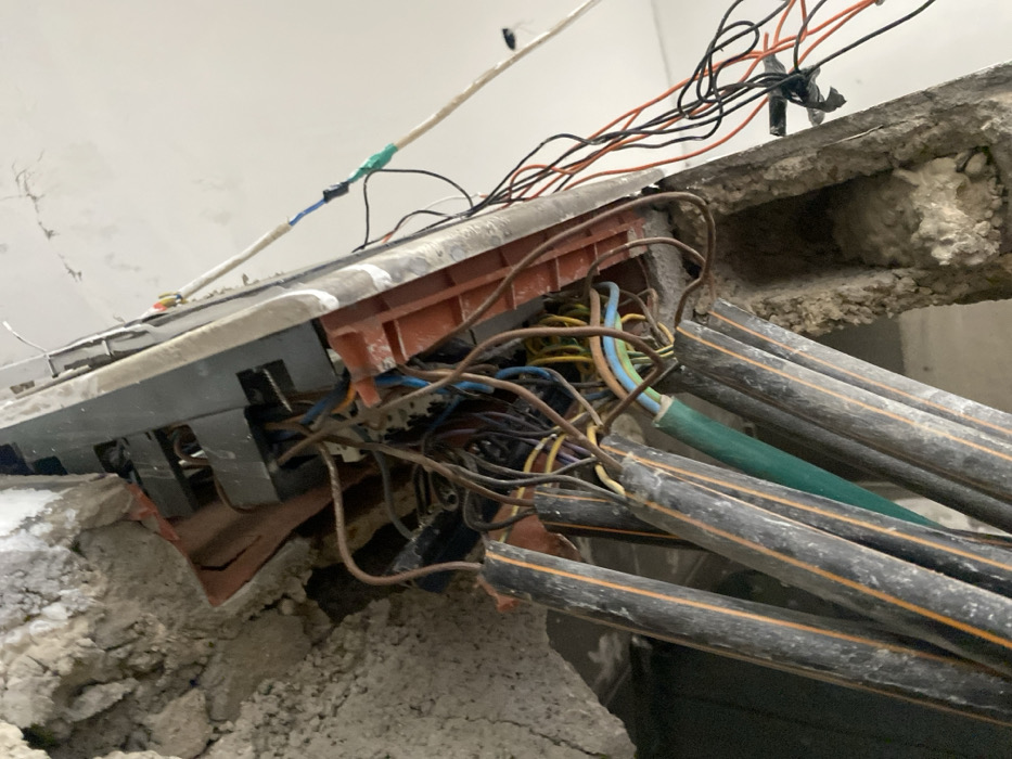 The image displays a close-up view of a chaotic arrangement of electrical wiring and conduits, partially embedded in a concrete or plaster wall. The scene suggests an exposed utility area, possibly due to demolition, construction, or disrepair.
In the center, a grey electrical box or distribution panel is visible, partially broken or pulled from its housing, revealing an intricate network of multicolored wires within. The box appears to be made of plastic, with some orange-brown internal components and grey external casing. Around the box, significant amounts of rough, crumbling concrete and plaster debris are present, suggesting an incomplete or damaged structure.
Numerous individual wires, varying in color (blue, yellow, brown, black, orange, green), emerge from and connect to the electrical box, spilling out in an untidy fashion. Several larger, dark grey cables, possibly conduits or heavy-duty electrical cables, with thin orange or yellow stripes running longitudinally, extend from the right side of the image towards the electrical box. One distinct green conduit or cable is also visible among these grey ones.
Towards the upper left, a white, seemingly insulated cable runs diagonally across the frame, connected to other wires with blue and green electrical tape, indicating a patched or temporary connection. Another cluster of thin, black and orange wires extends upwards from the top edge of the electrical box area. Black electrical tape is also seen securing some connections on these wires.
The background shows a plain, light-colored wall or ceiling, exhibiting some cracks and an uneven texture, further contributing to the impression of an unfinished or damaged environment. There is no clear indication of the time of day, but the lighting suggests an indoor setting. No people or discernible text are visible in the image. The overall impression is one of disarray and potential electrical hazard due to the exposed and disorganized wiring.