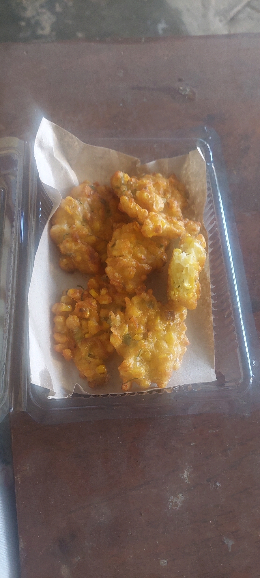 A close-up overhead shot shows a clear plastic container filled with golden-brown fried corn fritters. The fritters are arranged on a piece of light brown parchment paper within the container. Some of the fritters are partially broken, revealing a soft, yellow interior with visible corn kernels and green herbs. The container is placed on a dark, wooden surface that appears to be a table. The background is blurred, suggesting the image was taken indoors, possibly in a home or a local food stall in Lumajang, East Java, Indonesia.
