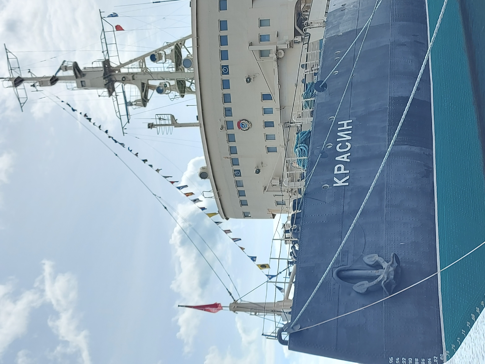 This is a photograph of the Russian icebreaker Krasin, a museum ship located in Saint Petersburg, Russia. The ship's name "КРАСИН" (KRASIN) is prominently displayed in white Cyrillic letters along its blue hull. An anchor is affixed to the hull. Depth markings are visible near the waterline. The ship's upper structure is a light cream color and has rows of windows. A mast and a complex arrangement of rigging are visible, with small flags strung between the masts. The sky is light blue with scattered white clouds.