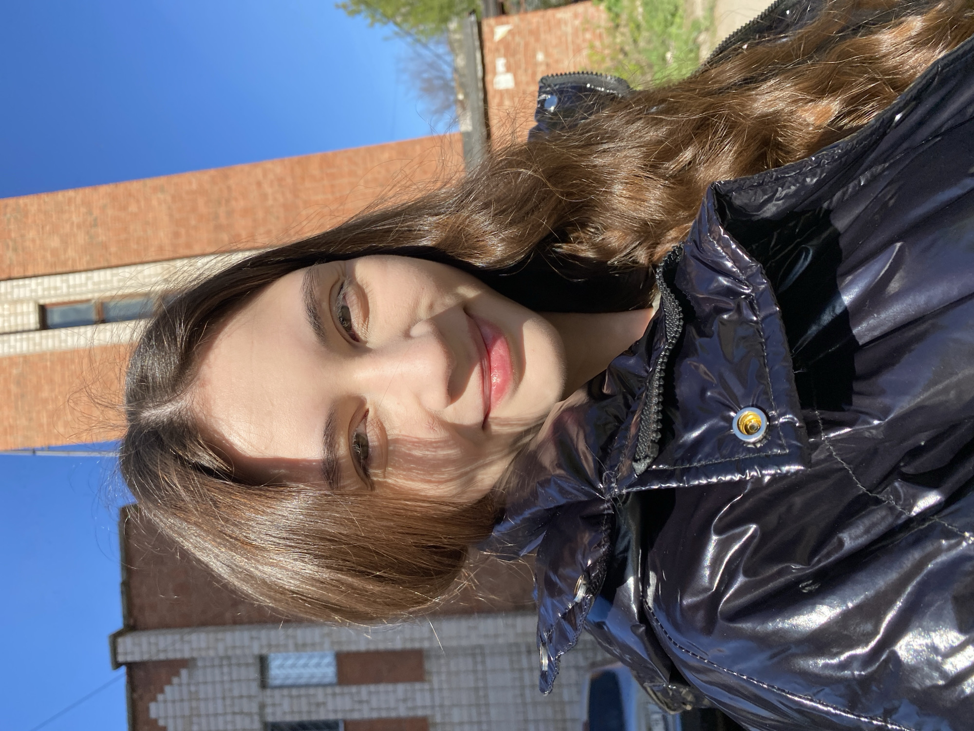 The image is a selfie taken outdoors. The subject is a young woman with long, wavy brown hair. She is wearing a shiny, black puffer jacket. A building, likely made of brick, is visible in the background, along with a clear blue sky and a glimpse of green foliage. There are no details that definitively pinpoint the location as Votkinsk, Russia.