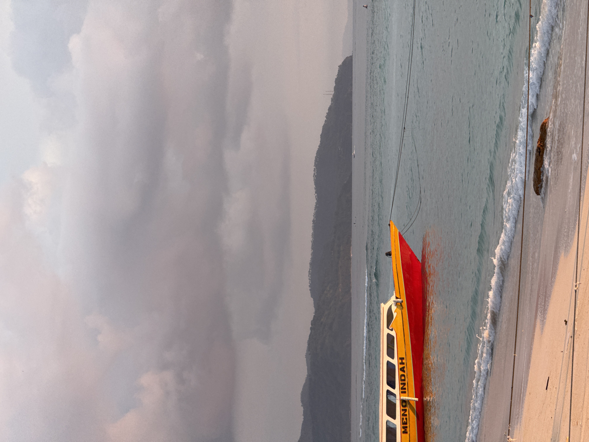 A medium shot, taken outdoors, features a red and yellow boat partially submerged in the sea, resting on a sandy beach. The boat has the words "MENO INDAH" printed on its side. The sea is calm with small waves, and a distant mountain or landmass is visible in the background, under a cloudy sky. The location is Unknown, Indonesia.