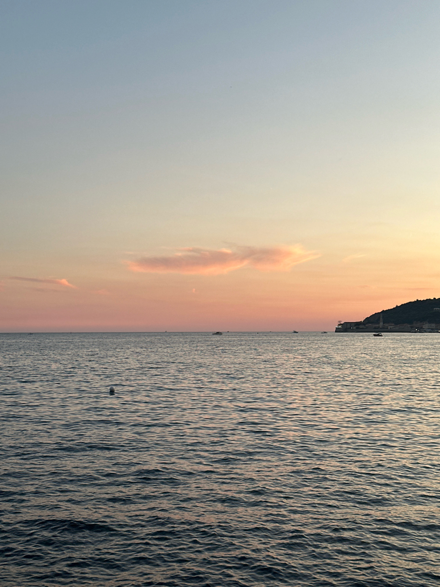 The image depicts a serene seascape at dusk, likely near Budva, Montenegro. The sky transitions from a pale blue at the top to soft hues of pink and orange near the horizon, indicating the sunset. A few wispy, pink clouds are scattered across the sky. The sea is calm, with gentle ripples reflecting the colors of the sky. Several small boats are visible on the water, some in the distance. In the far right of the image, a dark, tree-covered hill rises from the sea, with structures of a town visible on its slopes and a prominent tower-like building near the water's edge. A white buoy is floating in the foreground of the sea. The overall atmosphere is peaceful and tranquil.