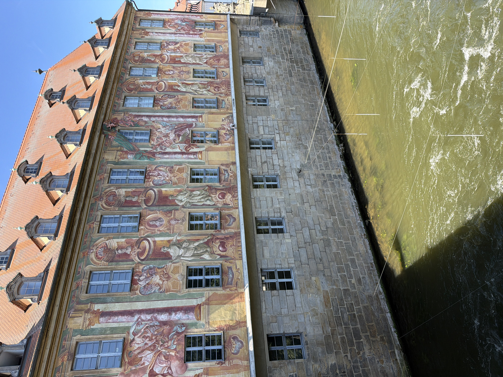The image shows a section of the Altes Rathaus (Old Town Hall) in Bamberg, Germany. The building is situated on an island in the Regnitz River, and the lower portion of the structure is visible, built of rough stone. Above this stone foundation, the building features a brightly painted facade with elaborate frescoes depicting mythological scenes and figures. A series of regularly spaced windows with blue frames are visible on both the stone and frescoed sections of the building. The Regnitz River flows in front of the building, its surface rippling and showing a greenish-brown hue. Several thin, white vertical poles, some connected by white string, are suspended above the water, creating a grid-like pattern. The sky above is a clear, bright blue.
