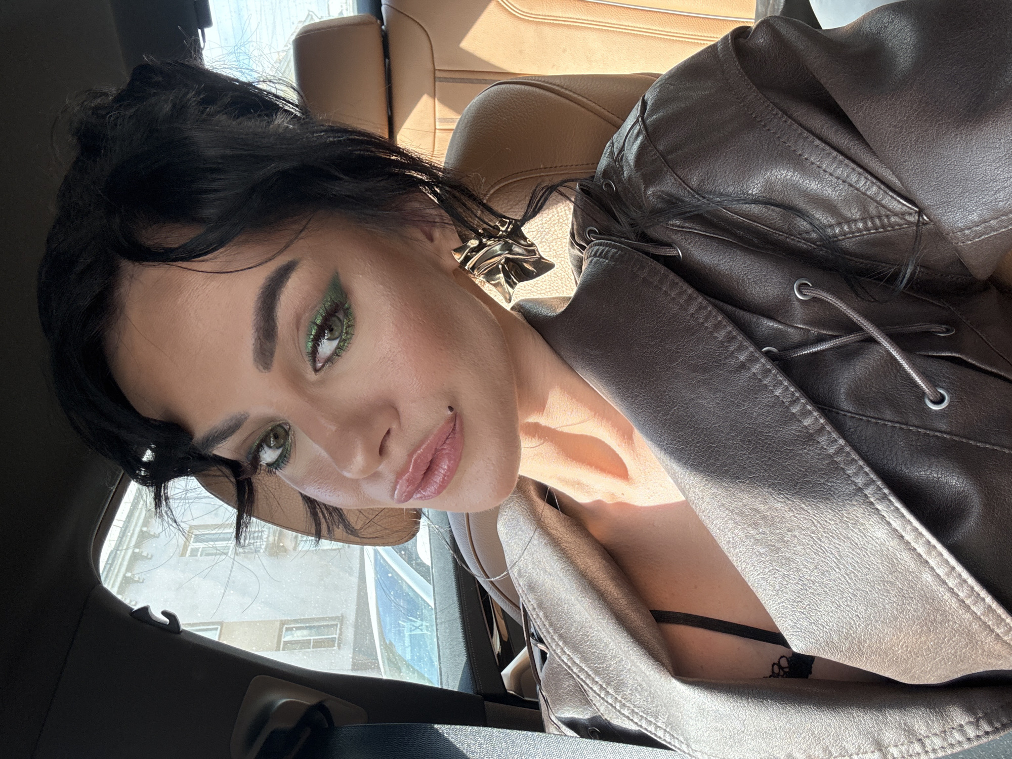 A woman takes a close-up selfie from inside a car during the daytime. She is positioned in the passenger or back seat, looking directly into the camera with a confident expression. Her dark hair is styled, and she wears prominent makeup, including bold green glitter eyeshadow, winged eyeliner, and pink lip gloss. A small, dark mole is visible above the right corner of her mouth.
She is wearing a textured, dark brown, possibly faux leather jacket with lacing details and grommets on the front, and a black bra strap is visible beneath it. A large, intricate silver earring adorns her right ear.
The car interior features light brown leather seats and dark grey trim. Strong, bright sunlight streams through the window, casting harsh shadows on her face and the car's upholstery, indicating a sunny day. Outside the car window, a light-colored building with multiple windows is visible, suggesting an urban or suburban setting. There is no visible text in the image. The location context for the image is Krasnoyarsk, Russia.