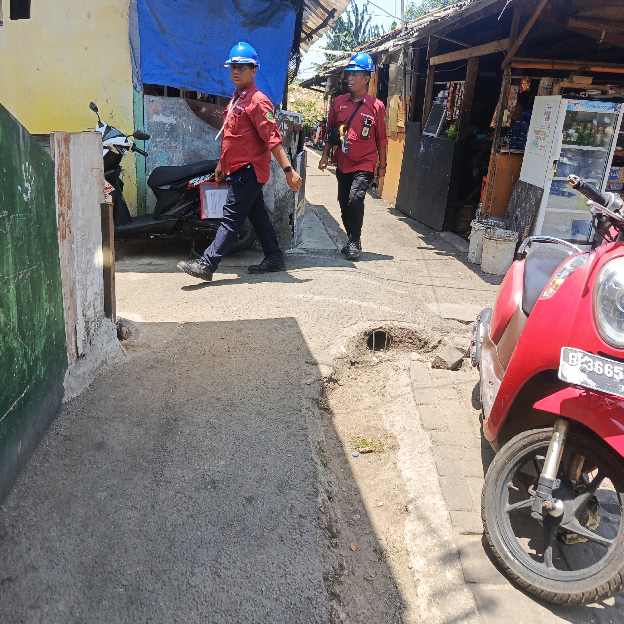 Two men wearing red shirts and blue hard hats are walking down a narrow alleyway. They appear to be inspectors or workers, as one is carrying a clipboard. The alley is lined with buildings that look like small shops or residences, with corrugated metal roofs and weathered walls. A red scooter is parked on the right side of the alley. The ground is a mix of asphalt and cobblestones. The scene suggests a densely populated urban area in South Jakarta, Indonesia, likely a commercial or residential neighborhood.