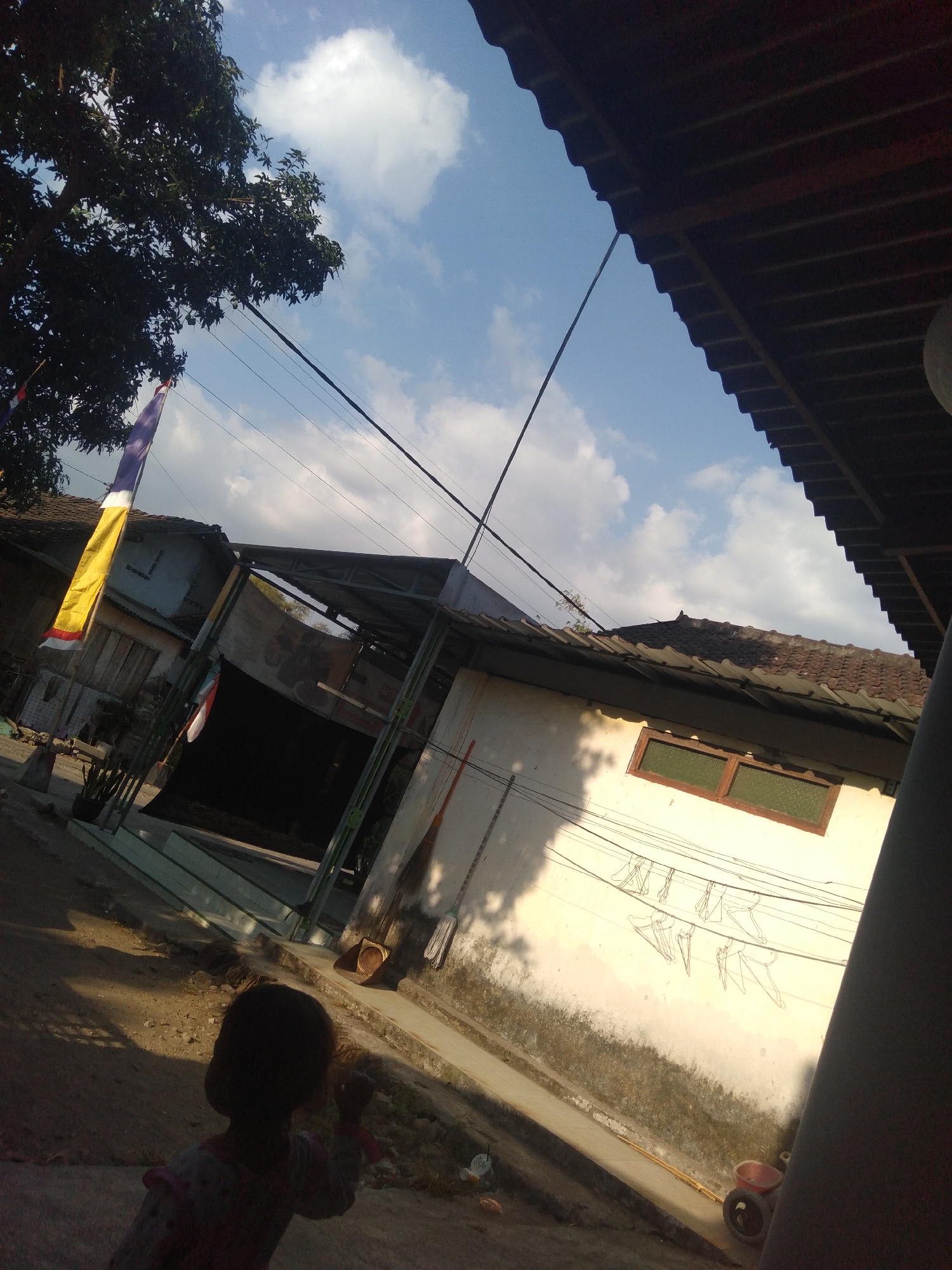 The image depicts a view of a residential area in Badas, Indonesia, with a tilted perspective. In the foreground, a child is partially visible, facing away from the camera. The background features several buildings with tiled roofs and a large tree with dark green foliage. Electrical wires crisscross the sky, and a partially unfurled yellow and red flag hangs from a pole near the tree. The buildings appear to be simple houses with some exhibiting signs of wear. The overall impression is of an outdoor scene in a somewhat rural or suburban setting during daylight hours, with a bright sky and scattered clouds. There are shadows cast by the structures and foliage, indicating sunlight. A broom and dustpan are visible near one of the buildings, suggesting everyday outdoor activities.