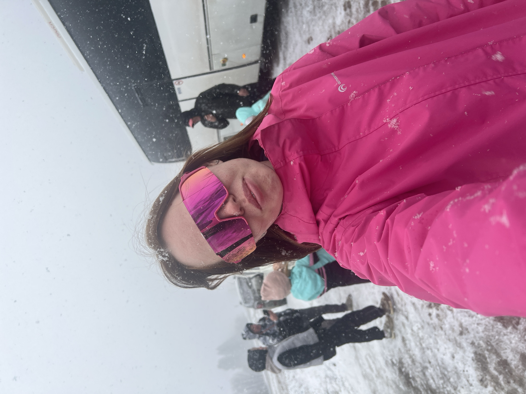The image shows a person wearing a pink ski jacket and pink-mirrored ski goggles taking a selfie in a snowy environment. In the background, other people are standing and some luggage is visible, suggesting a travel or outdoor setting. A large bus is partially visible on the left side of the frame. Snowflakes are falling. The location is Kliuchi, Russia, judging by the context and other images with similar composition.