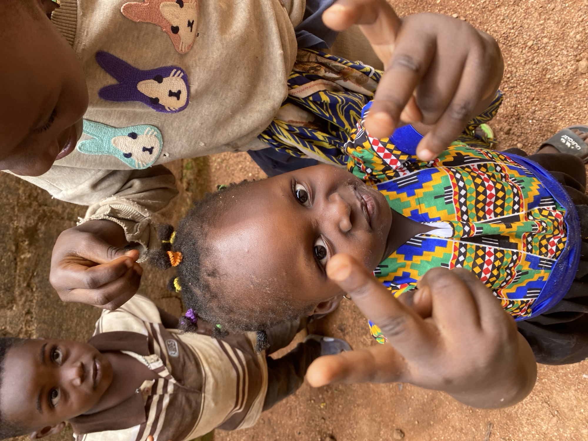 An overhead outdoor shot captures three young children from Kuru Dungus, Nigeria, positioned on reddish-brown dirt. In the foreground and center, a young child, likely a toddler, looks directly at the camera with a curious expression. Their hands are raised and appear to be gesturing towards the lens. They are wearing a vibrantly patterned top with a geometric design in yellow, blue, green, red, black, and white. Their dark hair is adorned with small, colorful ties in shades of orange, yellow, and purple.
To the upper left, an older child is partially visible, their face turned downwards. They wear a light-colored sweatshirt embellished with three rabbit appliques in pink, purple, and light blue. To the lower left, another child, also young, looks up at the camera with a slight smile, dressed in a brown and white striped polo shirt. A black sandal with the text "Adidas" in white is visible on the ground near the central child. The image is taken in natural daylight, suggesting a clear day, and captures a candid, interactive moment between the children and the camera.