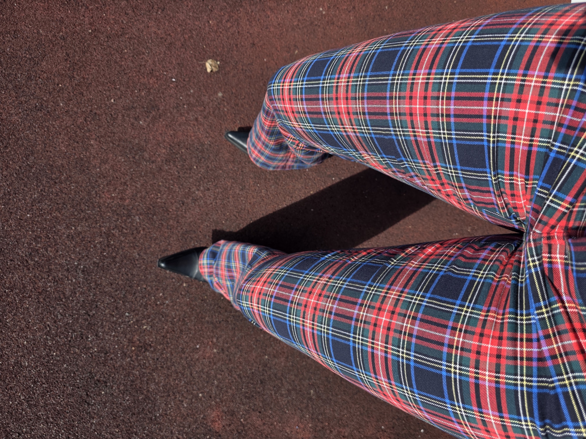 A low-angle, overhead shot shows the legs of a person wearing black pointed-toe shoes and red, blue, green, and yellow plaid pants. The person is sitting or standing on a coarse, reddish-brown surface, likely an outdoor recreational area or pavement. The lighting suggests it is daytime, with a shadow cast by the person's left leg towards the left side of the frame. A single dried leaf is visible on the surface in the upper right quadrant of the image. There is no indication of the specific location within Saint Petersburg, Russia.