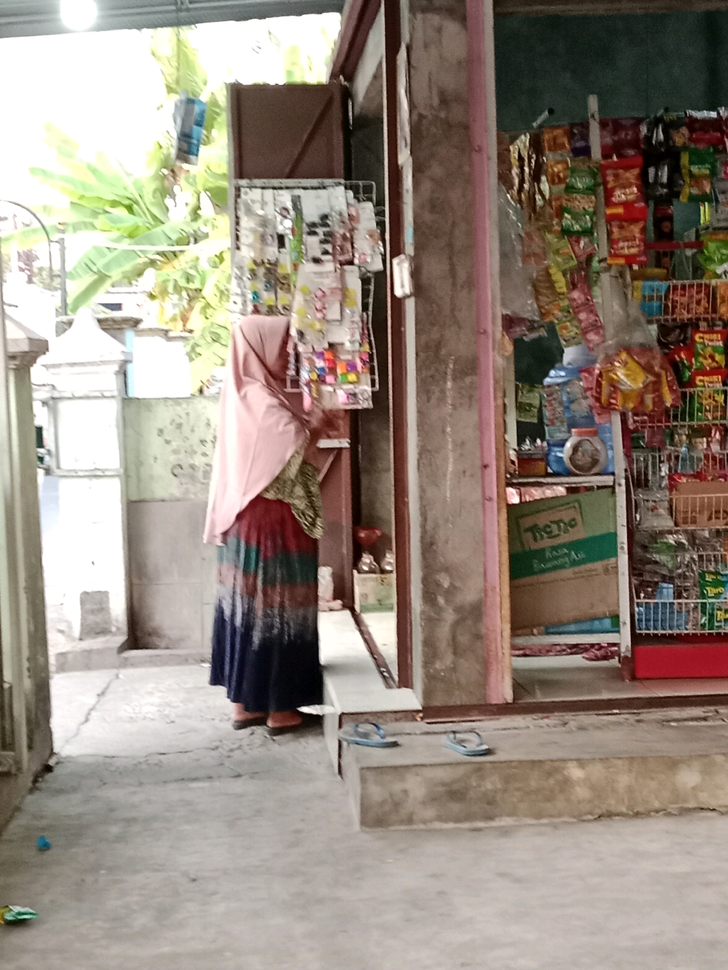 The image depicts a street-level view of a small convenience store in Waru, Indonesia. A woman wearing a pink hijab and a long, colorful skirt stands in front of the store, appearing to be browsing items displayed on a rack. The store itself is stocked with various packaged goods, including snacks and drinks, visible on shelves. A green cardboard box with text is also present. The ground is paved with concrete, and a pair of blue flip-flops are placed on a raised concrete step in front of the store. Banana trees are visible in the background, suggesting a tropical environment. The overall scene captures a moment of everyday commerce in a local Indonesian setting.