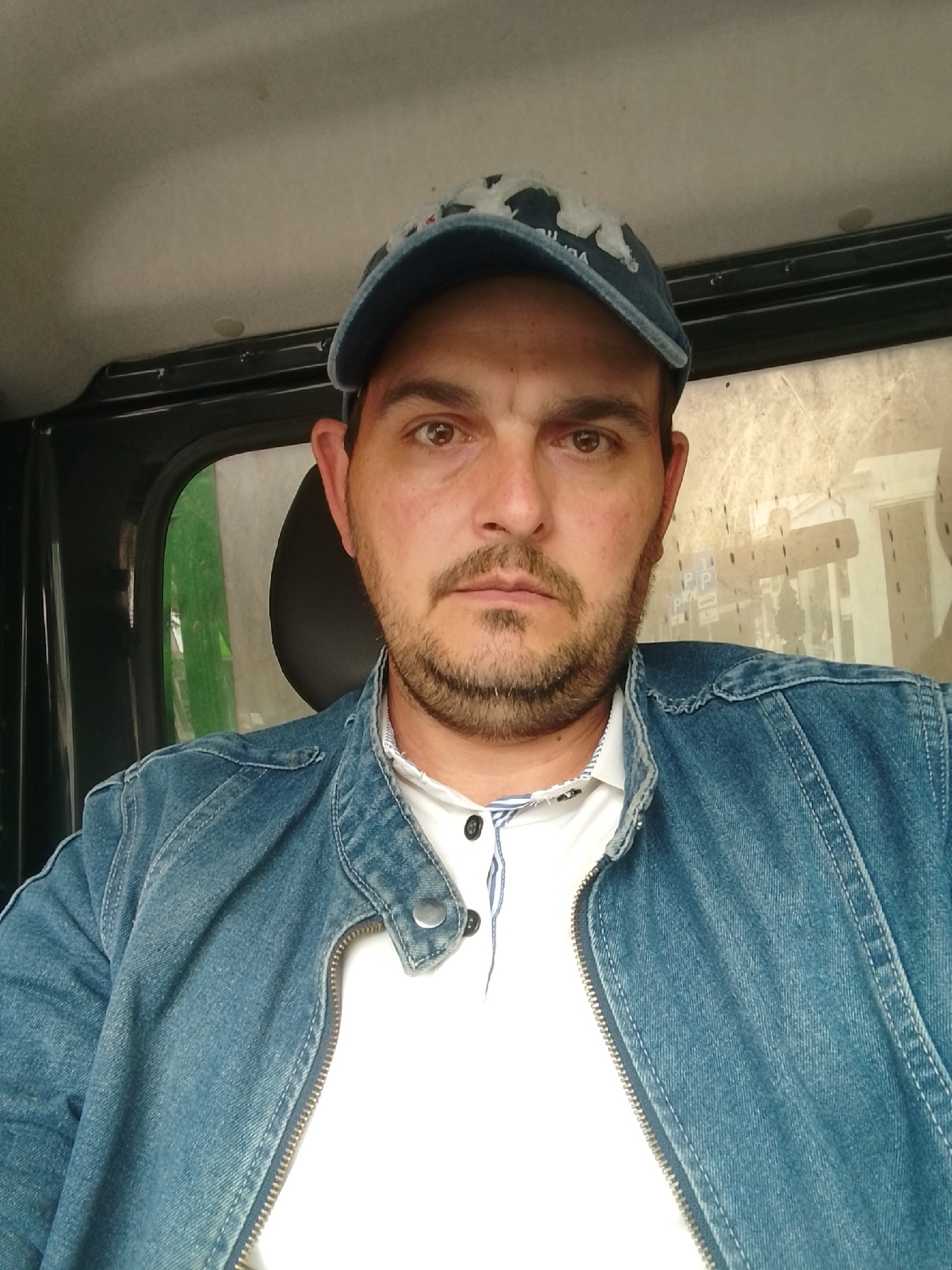 A man is shown from the chest up, sitting inside a vehicle. He is wearing a blue denim jacket over a white collared shirt with dark buttons. On his head, he wears a blue baseball cap with a white design on the front. The man has brown eyes, a beard, and is looking directly at the camera. The interior of the vehicle is visible, with the ceiling and a window in the background. The window shows a blurred outdoor scene with some structures and greenery. There is no indication that this image was taken in Pinneberg, Germany.