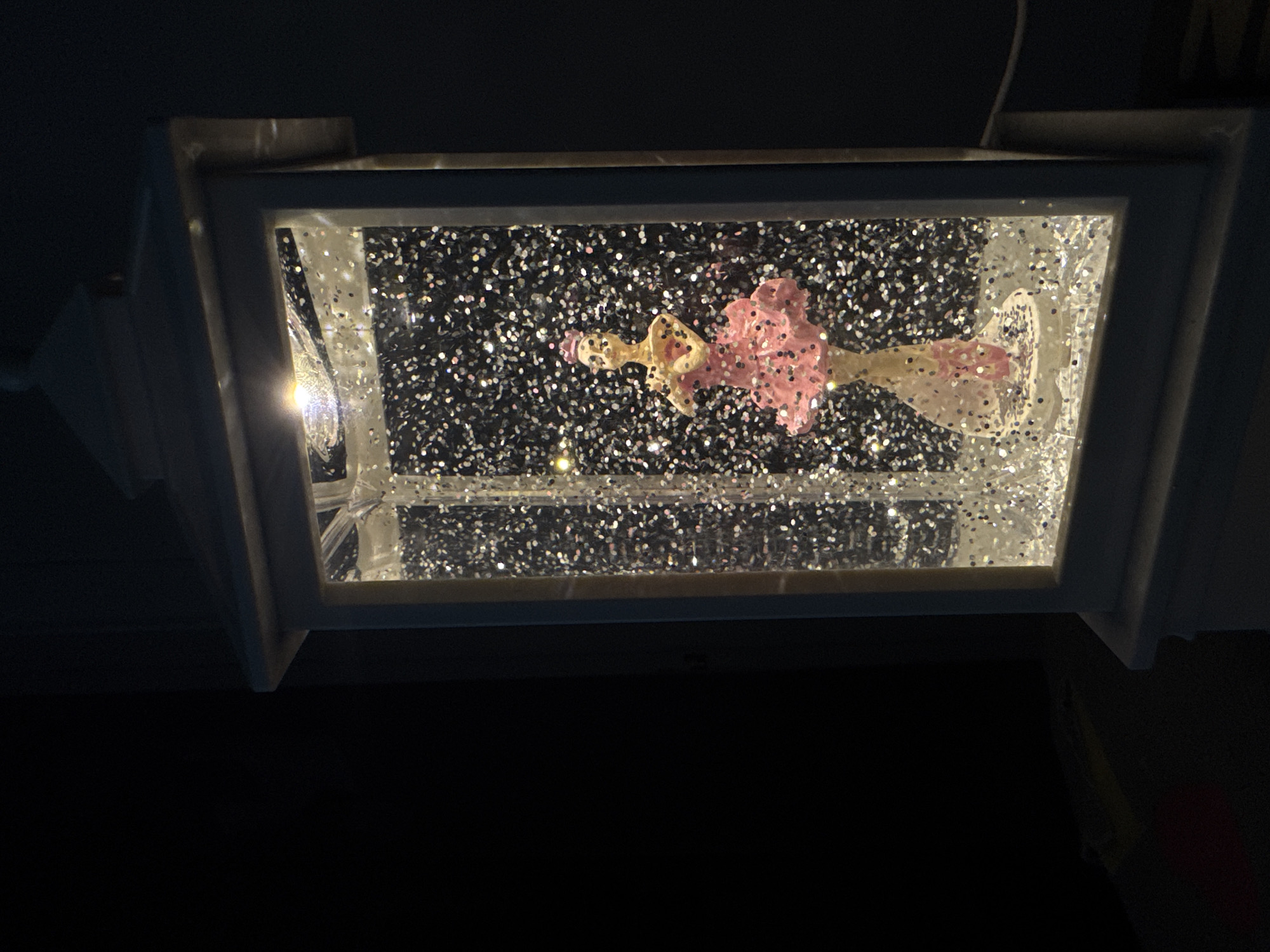 A low-angle, medium shot shows a snow globe enclosed in a white frame, potentially a decorative lamp fixture, against a dark background. The globe contains a figure, seemingly a ballerina with a pink tutu, suspended in a liquid filled with silver glitter particles. A light source illuminates the left side of the globe, causing a bright spot on the glass and highlighting the glitter. The image appears to be taken indoors, with the fixture mounted on a dark surface. The exact location within the city of Chaikovsky, Russia, cannot be determined from this image.