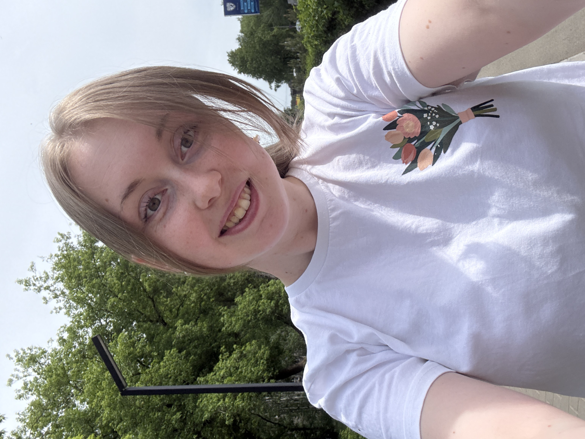 A young, fair-skinned woman poses for a selfie outdoors, on a sunny day. She has shoulder-length blonde hair, parted to the side. She is wearing a white t-shirt adorned with a floral bouquet design. Her head is slightly tilted to the side, and she is smiling, revealing her teeth. The background features a tree with green leaves and a section of an urban environment. In the upper left portion of the frame, there is a blue and white sign with text that is difficult to read. The image's location is the city of Unknown, Unknown.