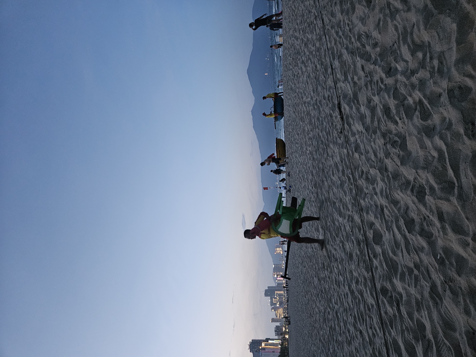 A lifeguard wearing a red and yellow uniform walks across a sandy beach in Đà Nẵng, Vietnam, carrying a green chair and a long pole. The city skyline, with illuminated buildings, is visible in the background under a pale blue twilight sky. Several people are scattered across the beach and in the shallow water, where two boats are present. Mountains are silhouetted against the horizon.