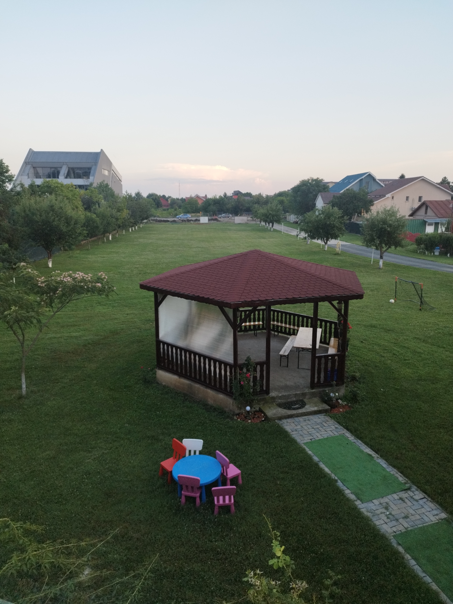 The image shows a residential area with houses and a large grassy lawn. A gazebo with a red roof is situated in the center of the lawn. In front of the gazebo, there is a children's table and chairs. A pathway made of pavers and artificial grass leads from the gazebo to the foreground. In the background, there is a modern building, houses, trees, and a street with cars. A small soccer goal with a ball is visible on the lawn to the right. The sky is overcast. The image depicts an outdoor scene in a suburban environment, likely a private yard or a park. The location is specified as Bucharest, Romania.