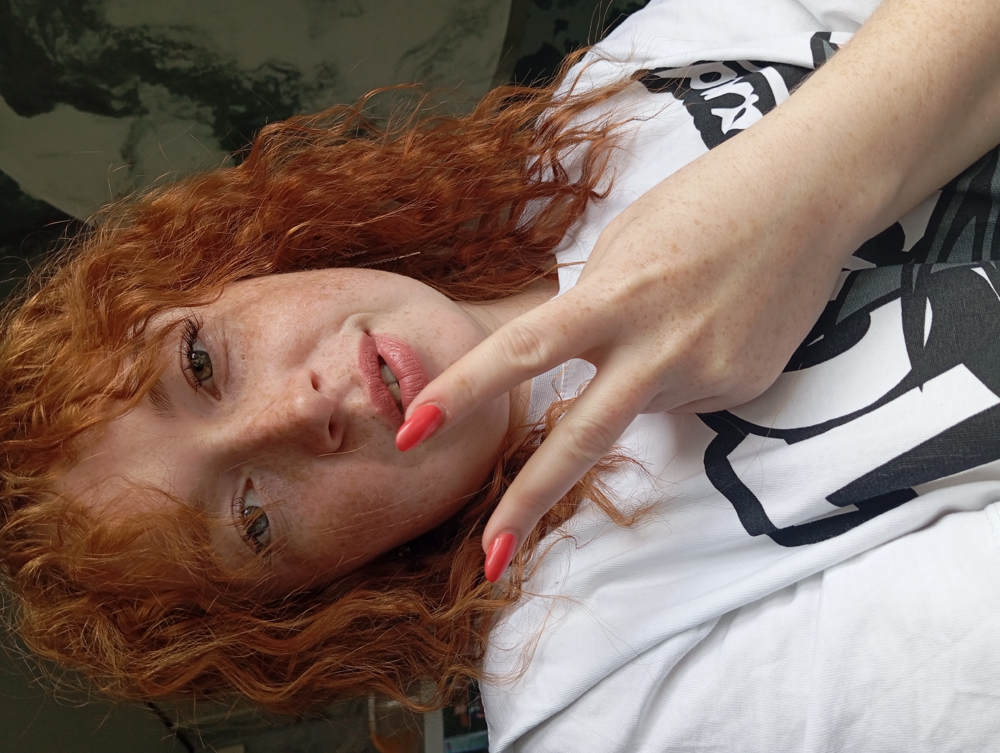 A close-up, tilted portrait shot features a woman with curly red hair and freckles. She is wearing a white t-shirt with a black and white design. Her left hand is raised, making a peace sign gesture. Her fingernails are painted a bright red color. The background is blurred and indistinct, revealing a dark, possibly marbled surface in the upper left corner. The photo's orientation is rotated counter-clockwise. No specific information suggests a connection to Yurgamysh, Russia.