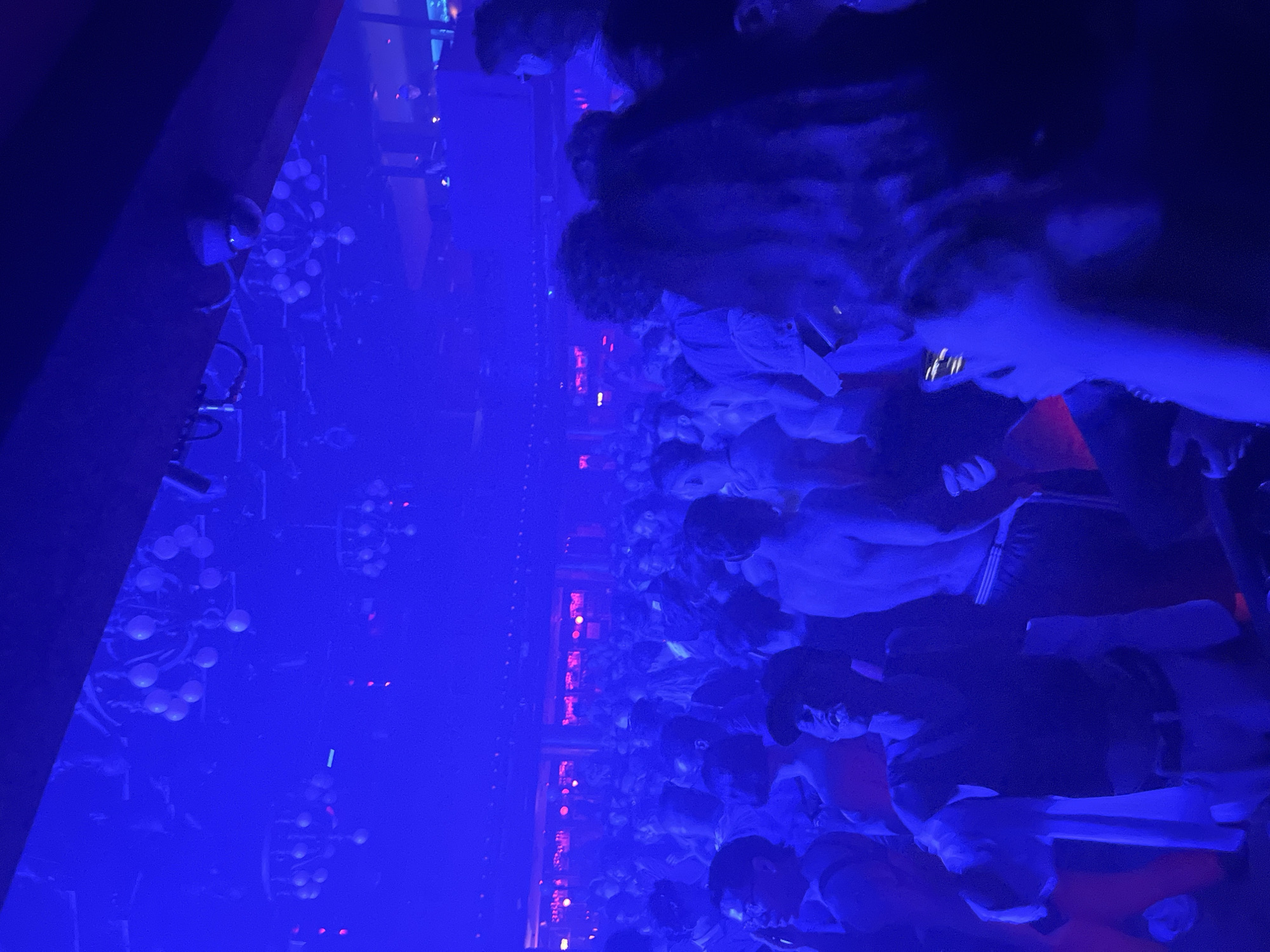 The image captures a dimly lit, crowded indoor scene, likely a nightclub or a party venue, bathed in predominantly blue light. Several chandeliers with spherical elements are visible suspended from the ceiling, adding to the atmospheric lighting. A dense crowd of people is packed together, with many individuals facing forward or looking upwards, suggesting they are engaged with a performance or DJ. Some people are holding up their phones, presumably to take pictures or record videos.
The people in the foreground are partially obscured by the low light and the density of the crowd, but some details are discernible. One individual in the center is wearing a dark baseball cap and appears to be looking upwards with a wide-mouthed expression. Another person to their left wears glasses. The overall atmosphere suggests a lively and energetic event. The location is specified as a nightclub in Barcelona, Spain.