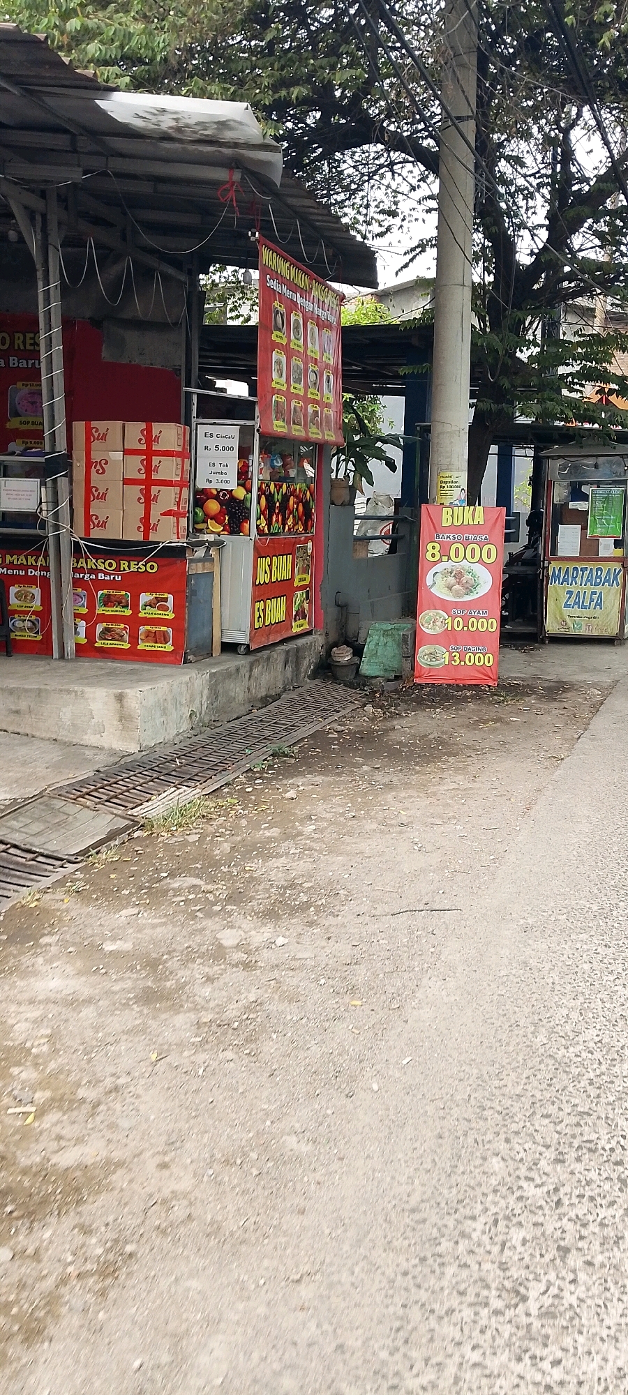 The image captures a street-level view of several food stalls in the city of Bekasi, Indonesia. The stalls are set up along a sidewalk, with a paved road visible in the foreground.
On the left, a stall displays signage that includes "BAKSO RESO MENU BARU" and images of food items. Boxes are stacked in front of this stall. Next to it, a fruit stall is visible with various fruits displayed, and signs advertising "ES CINCAL Rp 5.000" and "ES TEH JUMBO Rp 3.000". Further right, a red banner advertises "BUKA" with prices for "BAKSO BIASA 8.000", "SOP AYAM 10.000", and "SOP DAGING 13.000". To the far right, another stall has signage for "MARTABAK ZALFA".
The environment appears to be outdoors, with trees and utility poles visible in the background. The ground is a mixture of pavement and dirt, with a metal grate covering a drain. The lighting suggests it is daytime, possibly overcast or shaded by the trees. No people are clearly visible in the image. The overall scene depicts a typical street food vendor area in an urban Indonesian setting.