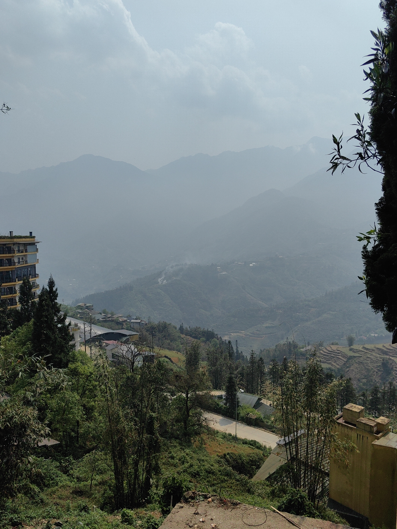 The image displays a mountainous landscape in Sa Pa, Vietnam, viewed under a hazy, light blue-grey sky with visible cloud formations. In the foreground on the left, a multi-story building with yellow exterior walls and dark-framed balconies is partially visible, flanked by dense green coniferous trees. A light-colored paved road winds through the mid-ground, passing several smaller structures with dark roofs nestled into the green, heavily vegetated slopes. Clumps of bamboo and other green foliage are prominent in the center-right foreground. A solid, light yellow/beige concrete structure with rectangular protrusions is partially visible in the lower right corner. The immediate foreground consists of disturbed earth with a piece of wood debris. In the mid-ground, particularly on the right, terraced fields are discernible, and wisps of smoke rise from a valley below. The background features multiple layers of mountains receding into a heavy atmospheric haze, appearing as faint silhouettes. No individuals or vehicles are present.