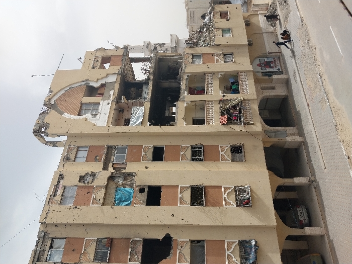 This image depicts a heavily damaged, multi-story residential building in Khan Yunis, Palestinian Territory, under an overcast sky. The building, rendered primarily in a light beige stucco with reddish-brown window surrounds and white decorative elements, shows extensive structural collapse on its upper floors, particularly towards the right side where a distinctive arched facade feature is partially destroyed. Numerous circular impact holes are visible across the building's facade, indicating widespread shrapnel or bullet damage. Sections of exterior walls are missing, revealing exposed interiors, including visible debris within rooms. Several windows are either shattered or covered with tarpaulins, one being distinctly blue and another white/silver. Balconies on the mid-levels display hanging laundry.
The ground floor features an arcade supported by arched columns, providing a sheltered area where a silver sedan is parked. A paved sidewalk runs along the building's base. On the sidewalk to the right, a person in dark clothing walks towards the right, while another individual is crouched beside a small group of children. A man in dark clothing walks past them towards the left. A large poster is affixed to the building's wall near the ground floor on the right. To the far right, beyond the main structure, the heavily damaged remnants of another building, possibly including a minaret, are visible amidst debris.