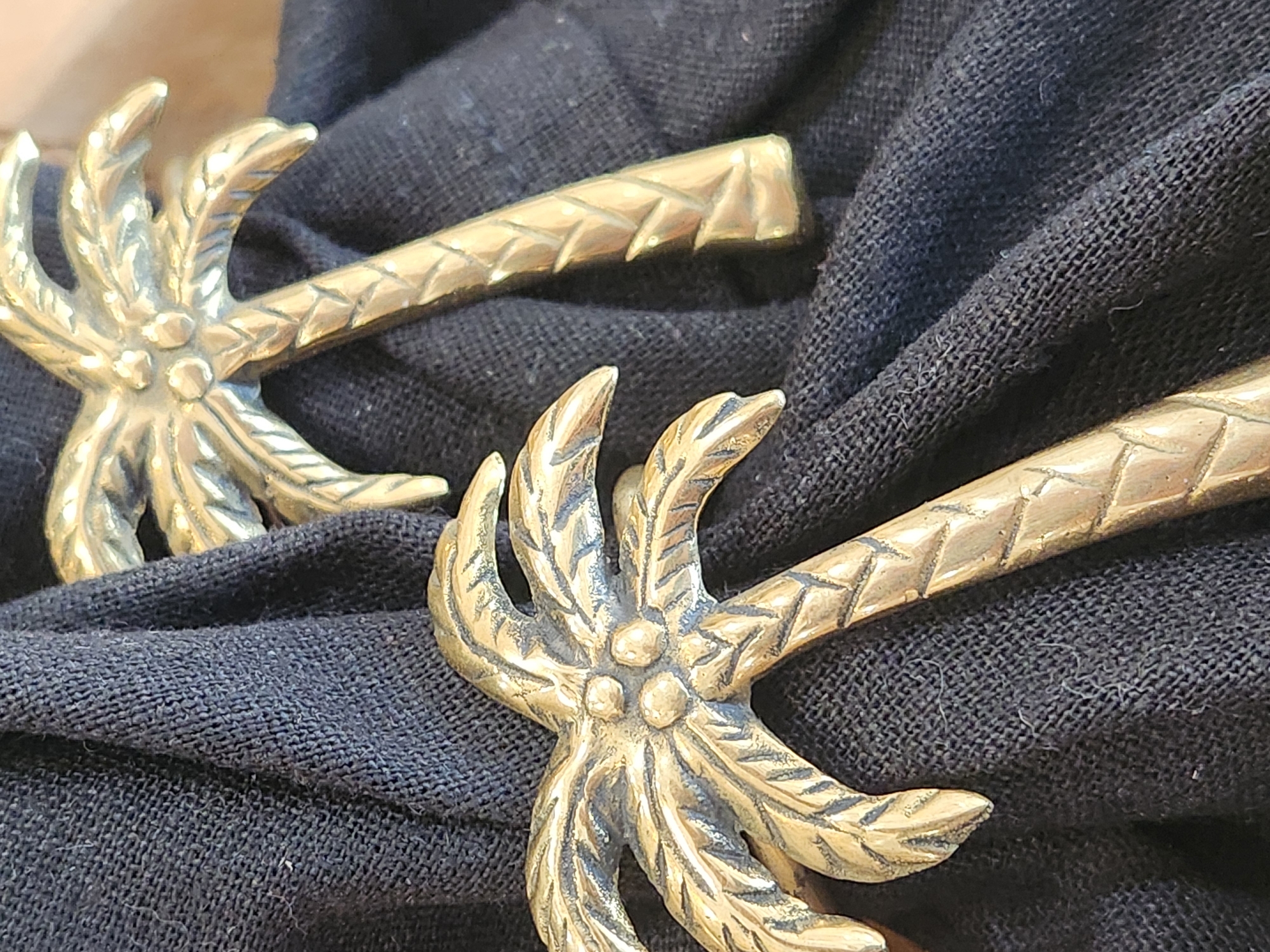 The image shows two brass palm tree-shaped objects resting on a dark, textured fabric. The objects appear to be decorative, possibly napkin rings or drawer pulls. The fabric is folded and bunched, creating shadows and highlighting the texture. The lighting is diffused, with a slight warm tone. The background is out of focus and appears to be a wooden surface. There are no people or animals in the image, and no obvious indications of location within Maitland, United States of America.