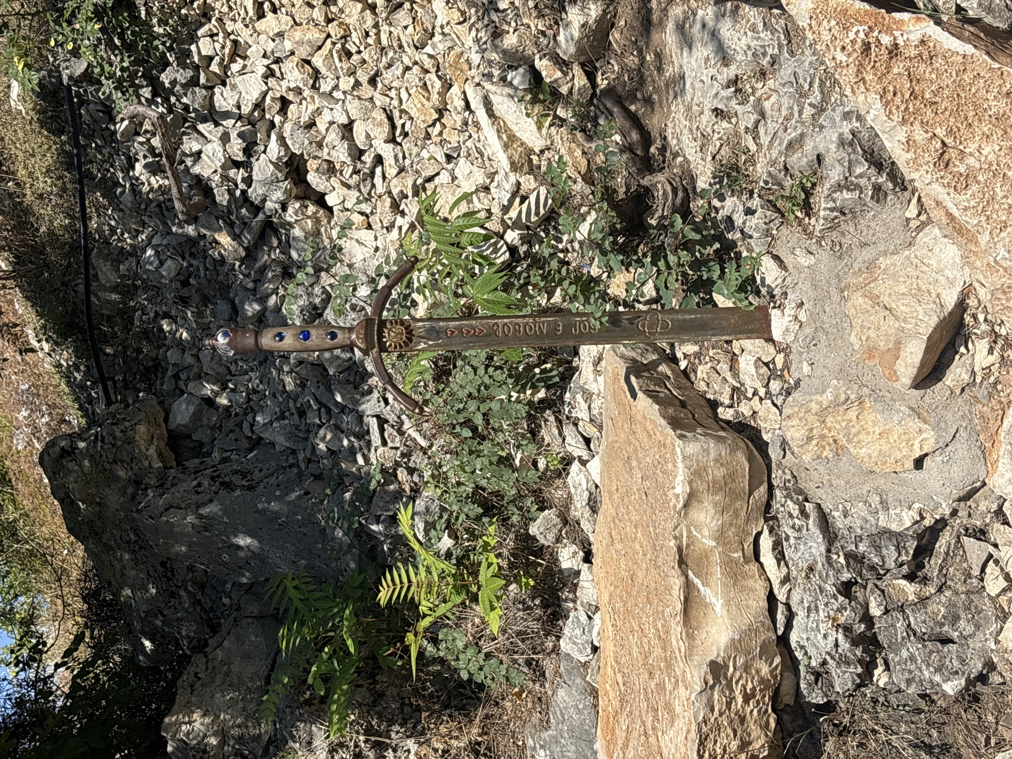 The image depicts a sword embedded in the ground amidst rocks and vegetation. The sword's hilt is adorned with blue gems and a sun-like emblem. Inscriptions are visible on the blade. The surrounding environment consists of a rocky slope with sparse greenery. The sword is situated near large, light-colored rocks. The scene is outdoors, under natural light. This appears to be a memorial or a tourist attraction, potentially related to local legends or history. The location is identified as the "Sword of Lukovit" in the city of Lukovit, Bulgaria.