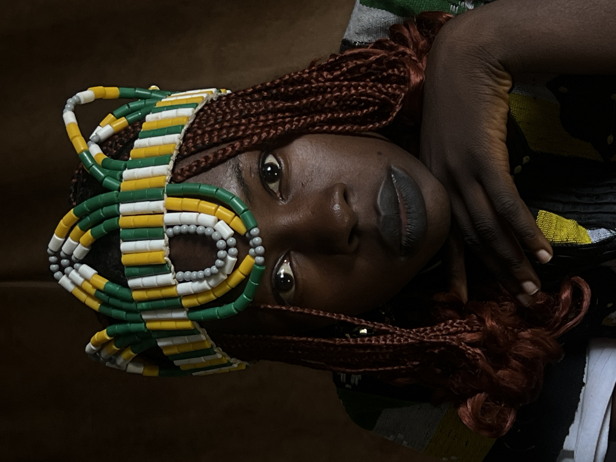 The image is a close-up portrait of a young woman with dark skin, wearing a vibrant, beaded crown. The crown is made of green, yellow, and white cylindrical beads arranged in a decorative pattern around her head. She has braided hair, a deep red-brown color, which falls around her face and shoulders. Her eyes are dark and appear to be looking directly at the camera. Her lips are painted with a dark, matte lipstick. She is resting her chin on her hands, which are clasped together. Her clothing is partially visible, showing a pattern of green, yellow, and grey stripes. The background is a plain, dark brown, suggesting a studio setting or a dimly lit environment. There are no identifiable landmarks or specific details that would pinpoint a location within the city of Jos, Nigeria. The image is focused on the subject's face and adornments, creating an intimate and regal feel.