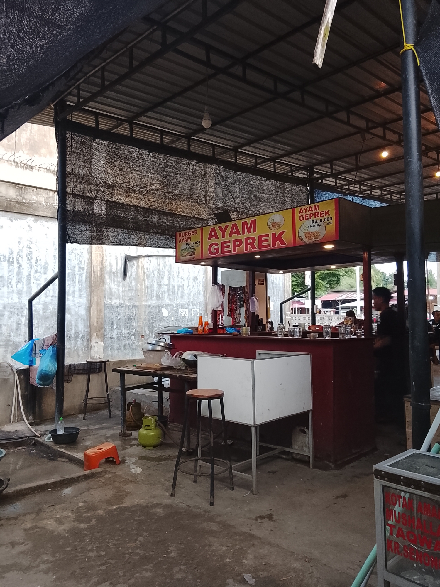 This image depicts an outdoor food stall in Lhokseumawe, Indonesia. The stall features a prominent yellow sign with red lettering that reads "AYAM GEPREK" (smashed chicken), with prices for "Rp. 8.000" and "Nasi Rp. 1.000". To the left of this sign, a smaller sign advertises "BURGER AYAM Rp. 12.000". The stall has a red counter with glasses and bottles on it. A white metal display case is situated in front of the counter, and to the left of that, a green gas cylinder is visible on the ground. A small orange plastic stool is also on the ground. In the background, a rough concrete wall with vertical lines is visible. Several people are present: a man in a black shirt is standing to the right of the stall, and two other individuals are seated further back in the stall area. The overall environment appears to be a busy, informal dining area.