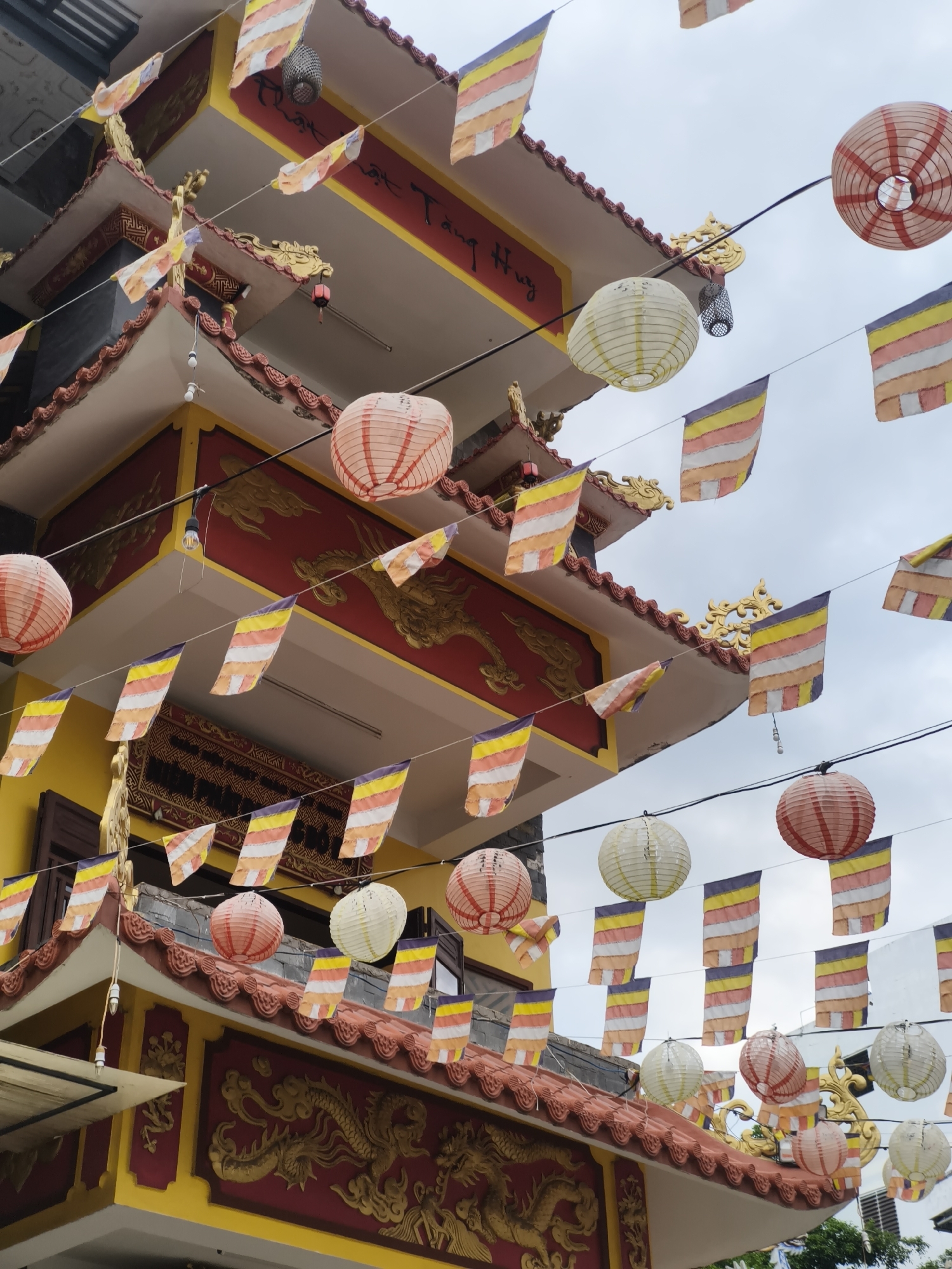 The image captures a close-up, low-angle view of a vibrant Buddhist temple facade in Nha Trang, Vietnam, adorned with numerous decorative elements. The temple itself is painted in bright yellow and red, with intricate gold dragon motifs embellishing its eaves and architectural details. Suspended on wires are strings of colorful prayer flags, each featuring horizontal stripes of yellow, orange, white, and purple. Interspersed with the flags are various paper lanterns in shades of red, white, and pink. The sky above is overcast and pale. There are no people visible in the frame. The image appears to be taken from street level, looking upwards at the temple's upper levels and rooflines.