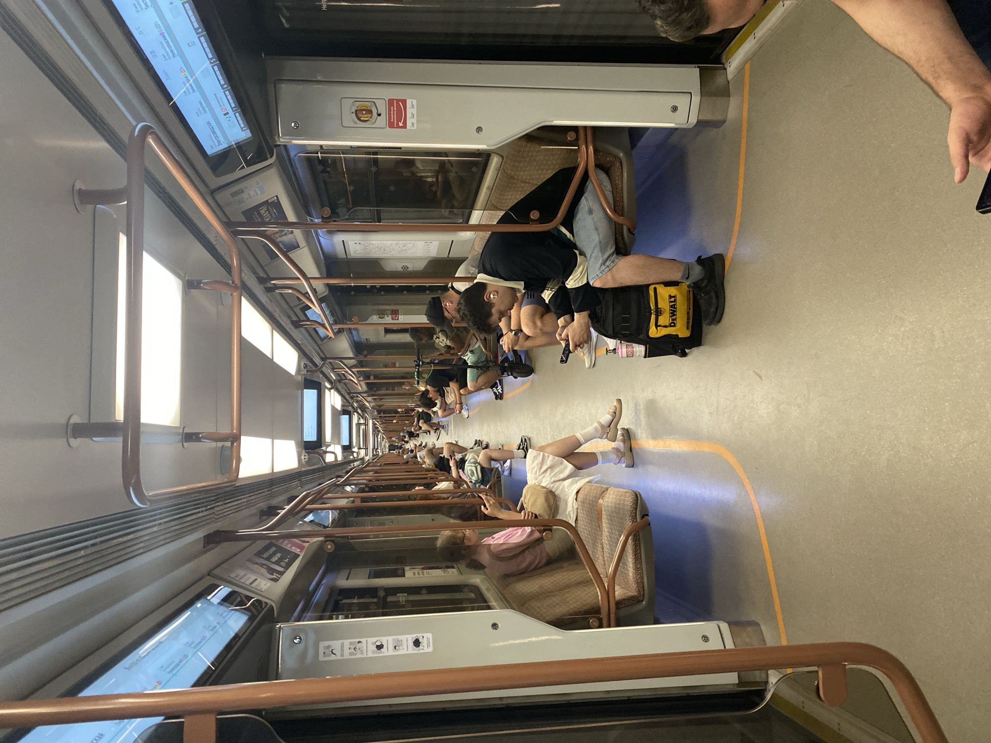 The image shows the interior of a subway train with several passengers seated and standing. The train has brown seats along the sides and a metal railing running along the ceiling. Digital screens are mounted above the windows, displaying information in a foreign language. The floor is a light gray color with an orange line marking a path. One woman is seated on the left, wearing a pink shirt and white skirt, looking at her phone. On the right, a man wearing a black and white striped shirt and blue shorts is seated, also looking at his phone, with a yellow DeWalt bag at his feet. Other passengers are visible in the background, some looking at their phones and others conversing. The lighting appears to be artificial, emanating from the ceiling panels. The overall environment suggests a public transportation setting within a city.