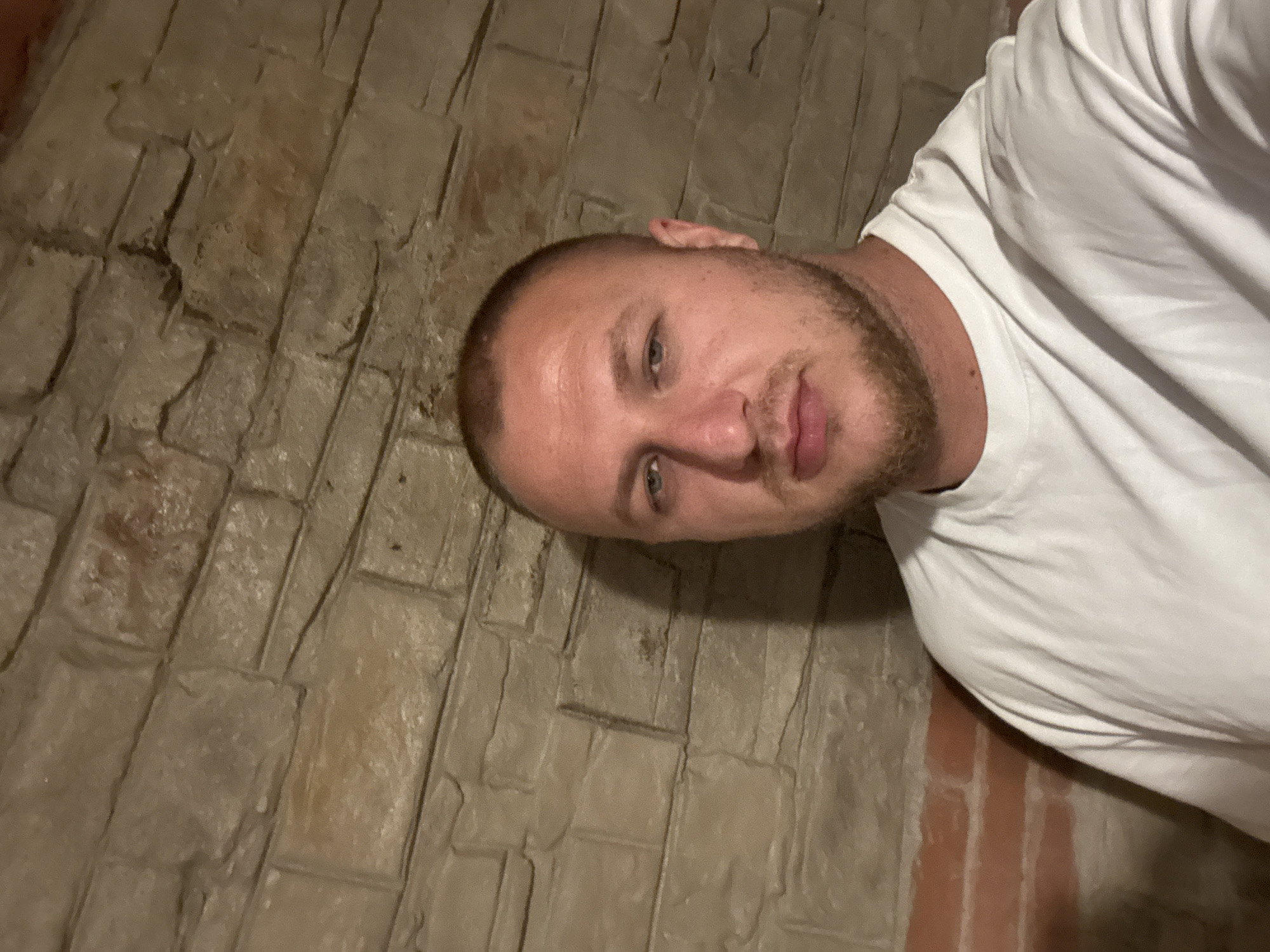 A rotated close-up portrait shows a man looking directly at the camera. He has short hair and a neatly trimmed beard. He is wearing a plain white t-shirt, which is visible on the right side of the frame. The man's face is positioned on the left, with his head tilted slightly. His expression is neutral.
The background features a textured wall constructed from irregular, light brown or grey stone blocks, possibly a type of brick or stacked stone. The wall has deep grout lines, giving it a rustic appearance. The lighting is dim and appears to be artificial, casting soft shadows that emphasize the rough texture of the wall and the man's facial features. The scene suggests an indoor setting or a sheltered outdoor space, possibly an old building or a basement. There is no visible text. Given the location context of Asenovgrad, Bulgaria, the stone wall could be characteristic of local traditional architecture.