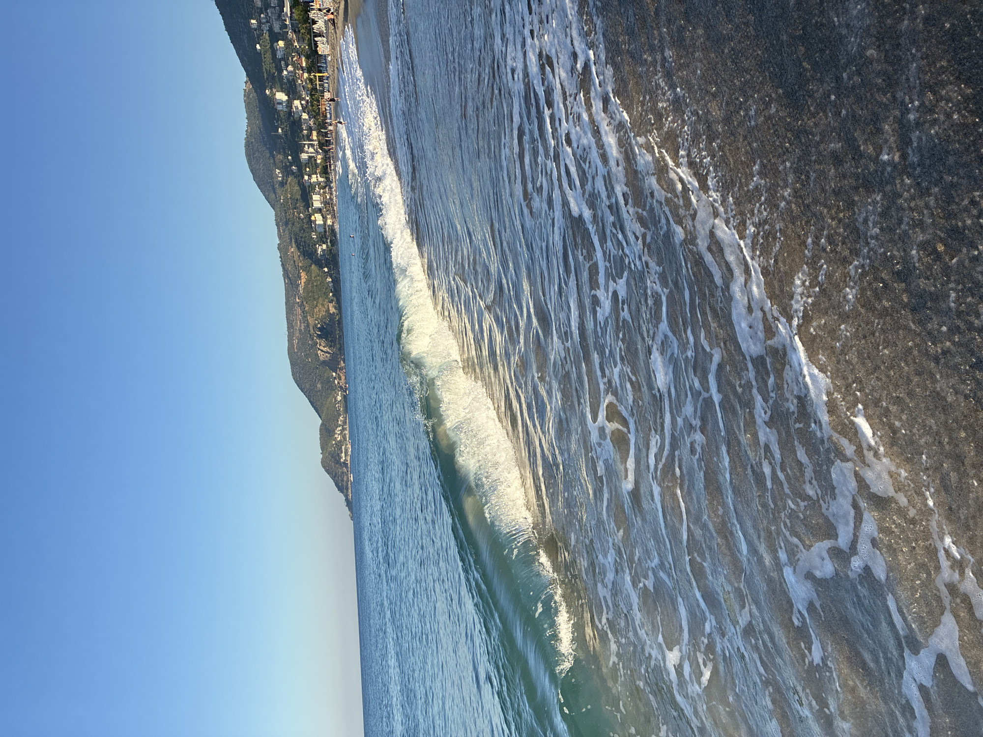 This is a photograph of a beach with a gently rolling wave breaking on the shore. In the background, a coastal town with buildings is visible nestled against a hillside under a clear blue sky. The water is a turquoise color, and the sand is dark. The scene suggests a serene and sunny day. While the specific location within Alanya, Turkey, cannot be determined from the image alone, it is representative of the city's coastal geography.
