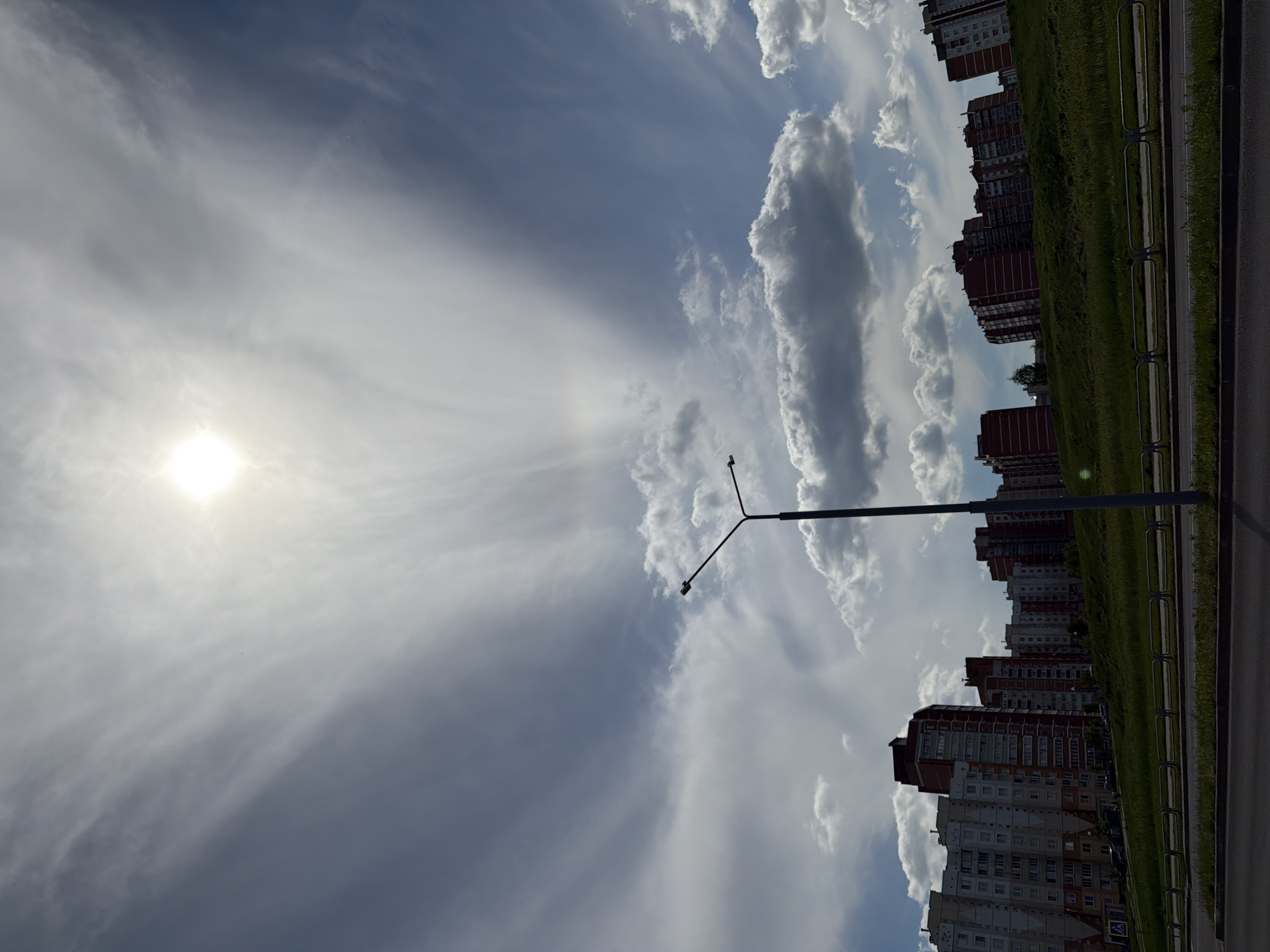 The image shows a landscape with a light pole in the foreground, extending into a cloudy sky with the sun visible on the left. To the right, there is a row of multi-story apartment buildings adjacent to a grassy area, followed by a road with a guardrail. The location is likely within a city.