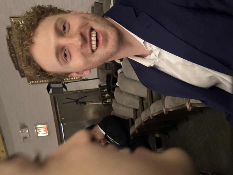 A man with curly blonde hair wearing a navy blue suit and a white shirt smiles towards the camera. He is standing in what appears to be a theatre or auditorium, as indicated by the rows of empty seats behind him. An exit sign can be seen on a wall, and another person wearing a black jacket is sitting in one of the seats. Part of a face is visible in the lower portion of the frame, potentially from the person taking the photograph.
