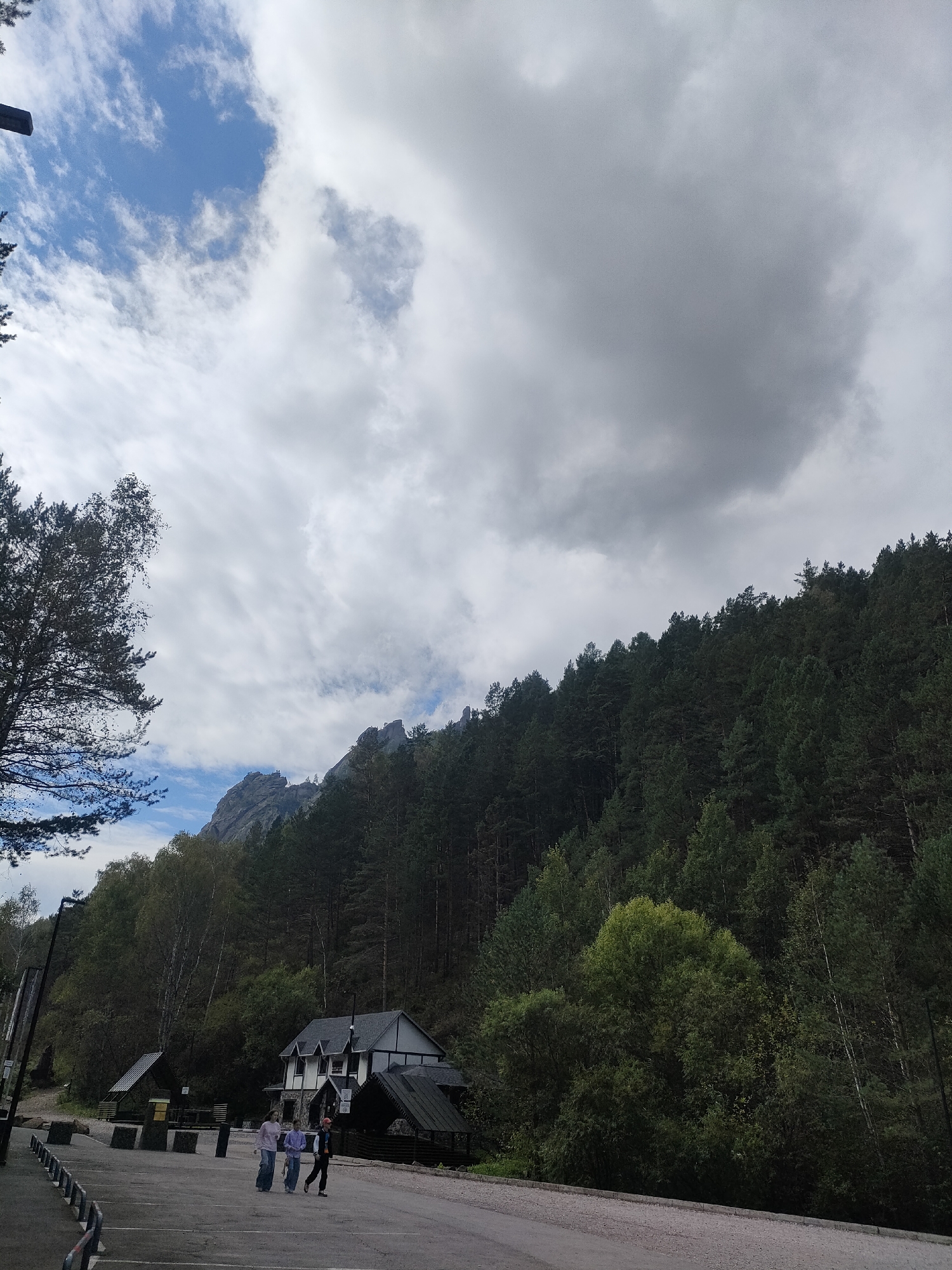 In an outdoor scene, a paved area in the foreground is partially obscured by a metal railing. Three individuals are walking across the paved area from left to right. To the left of the paved area, a building with a dark roof and white walls is visible. Behind this building, a dense forest covers a hillside that leads up to rocky mountain peaks under a cloudy sky. A tall tree with dark foliage stands on the left side of the image, framing the view of the mountains and sky.