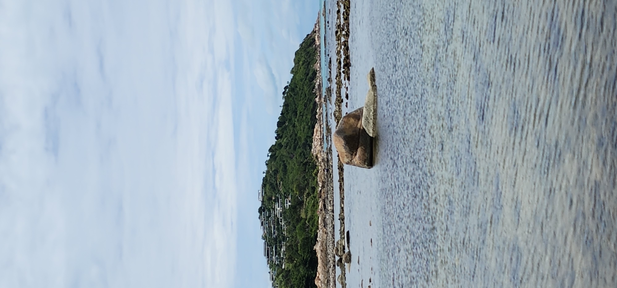 The image depicts a coastal scene on the island of Ko Samui, Thailand. In the foreground, calm, shallow water reflects the cloudy sky. Several large, rounded rocks are visible protruding from the water. In the mid-ground, a rocky shoreline lines the base of a lush, green hill. The hill is densely covered in trees and foliage, with numerous white buildings and structures clustered on its slopes, suggesting a developed resort or residential area. The horizon is dominated by a hazy sky with scattered clouds.