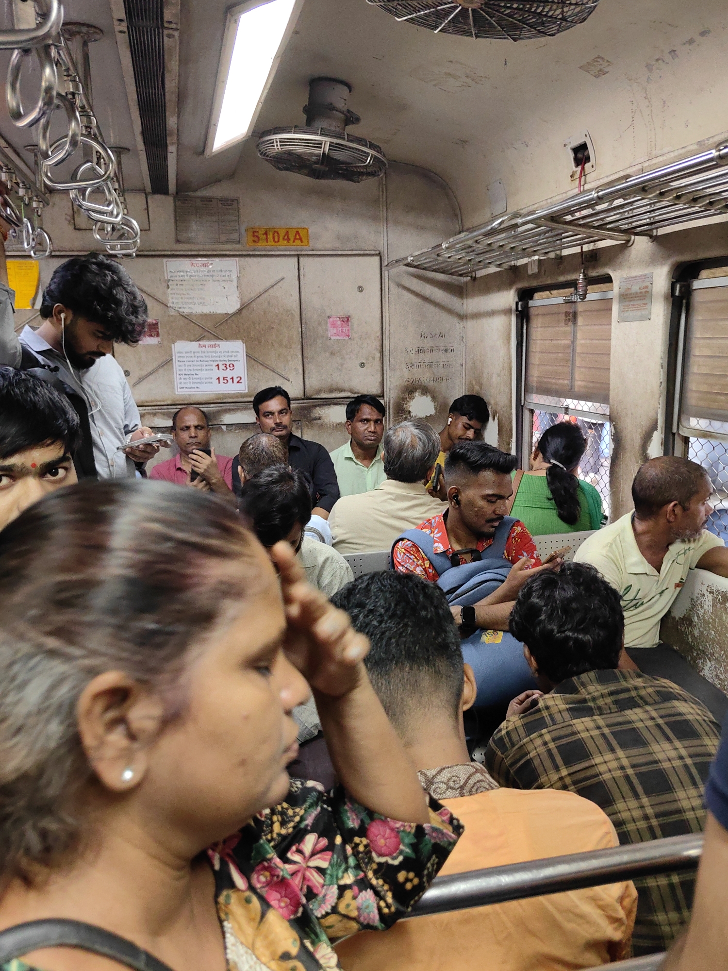 The image depicts the interior of a crowded Mumbai local train carriage during daytime. The scene is densely packed with passengers of various ages and genders, many of whom are seated and some standing, with the closest individuals blurred due to their proximity to the camera. The carriage walls and ceiling are worn and grimy, featuring signage, a ventilation fan, and metal grab handles. Passengers are dressed in casual attire. A man in the foreground on the left is partially visible, looking down, while a woman in the center foreground is out of focus, with her hand near her face. Other passengers are visible engaged in activities such as looking at their phones or conversing. The overall impression is one of everyday commute in a busy urban environment.