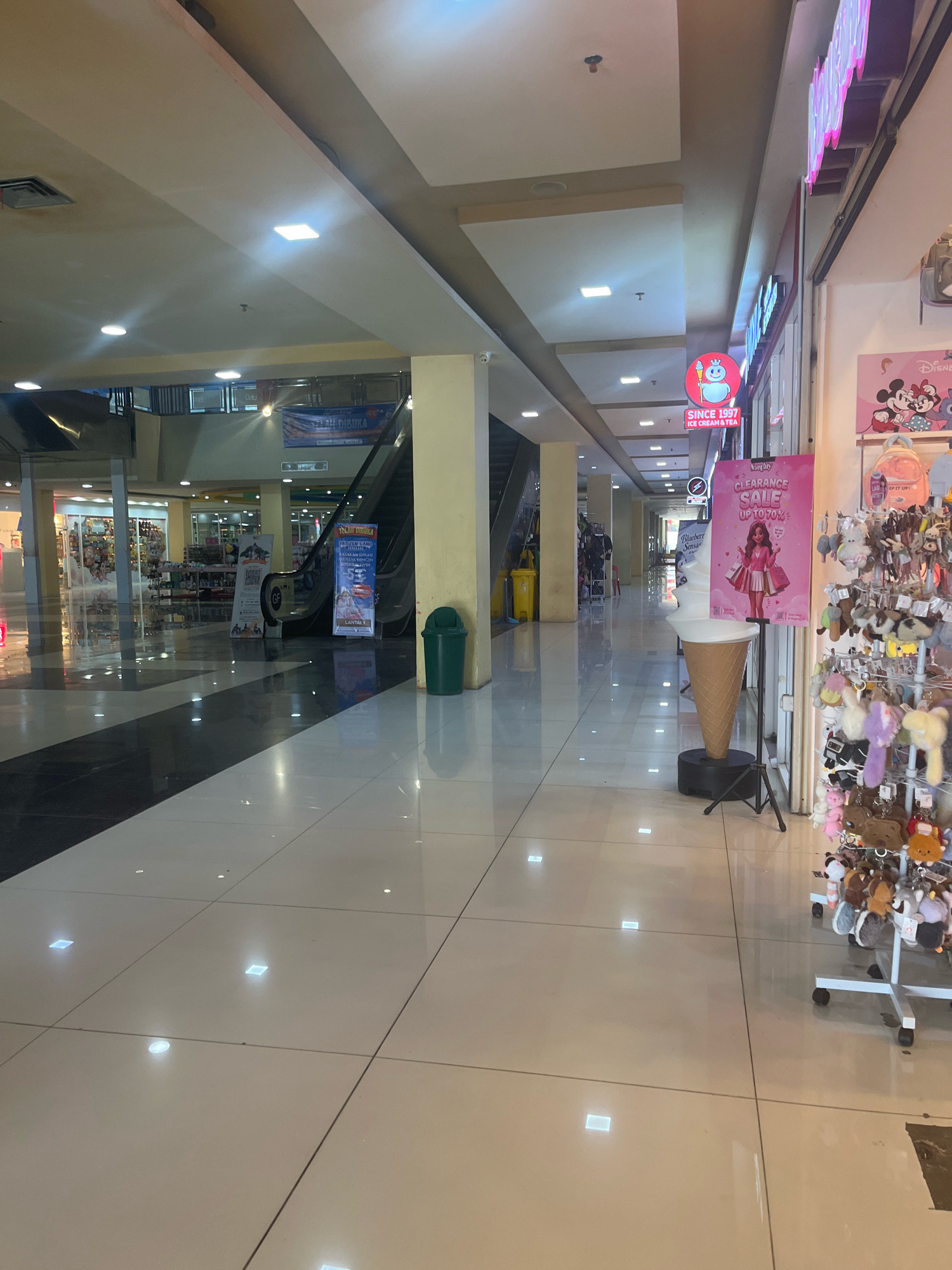 The image captures an indoor scene, likely a commercial establishment in Sengkang, Indonesia, as suggested by the presence of an "Ice Cream & Tea" sign with text that appears to be in Indonesian. The environment is a well-lit corridor with polished tile flooring that reflects the overhead lights.
On the left side of the corridor, there are shops with displays of merchandise, including shelves stocked with items. An escalator is visible in the background, leading to another level of the establishment. A green trash can stands near the escalator.
On the right side, a store display features numerous keychains and small plush toys hanging from a rack. A large, decorative ice cream cone stands on a stand, next to a pink promotional poster advertising a "Clearance Sale Up to 70%." Above this display, a neon sign with pink lettering is visible.
The lighting is bright, with recessed ceiling lights and some square illuminated panels integrated into the ceiling structure. The overall impression is of a clean and modern shopping area. No individuals are clearly depicted in the image.