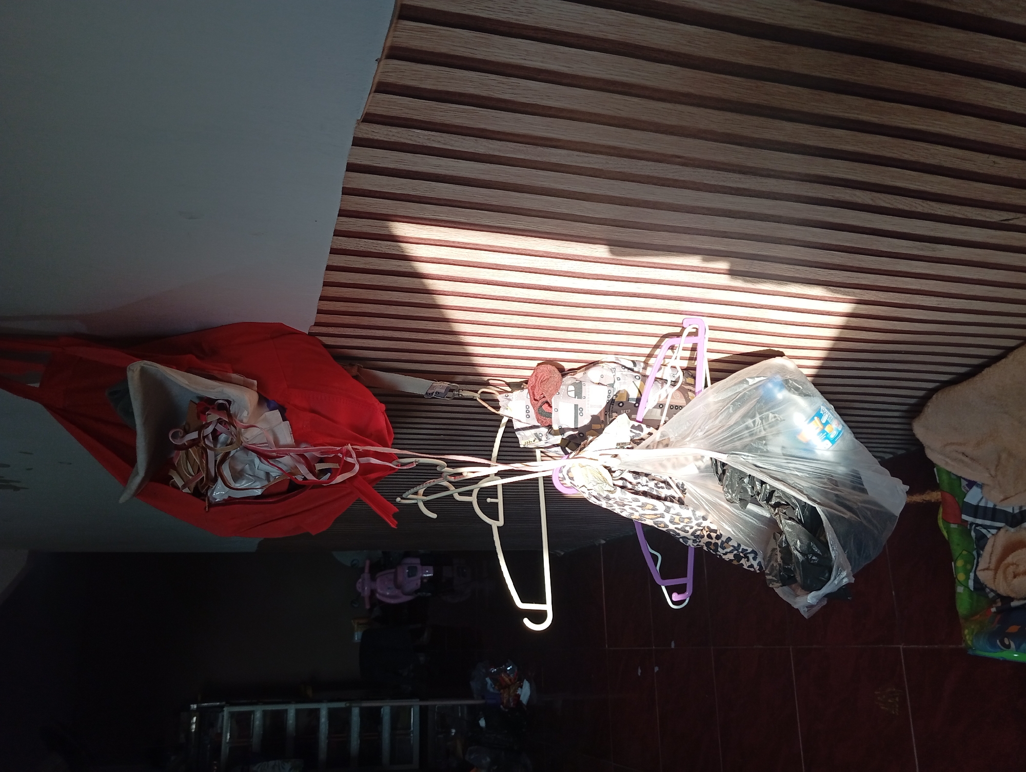 The image depicts an interior scene in Jogosari, Indonesia, likely a residential or storage area. A large red bag, filled with various colorful ribbons and fabrics, hangs from the upper right corner of the frame. Below it, several clothes hangers are suspended, holding a mix of items including a white plastic bag filled with dark objects, a patterned fabric, and another item with a car print. To the left, a metal shelving unit is visible in the dimly lit background, along with what appears to be a pink toy scooter. The wall behind the hanging items is covered with vertical wooden panels, with sunlight casting distinct shadows across them. The floor is tiled in a reddish-brown hue.