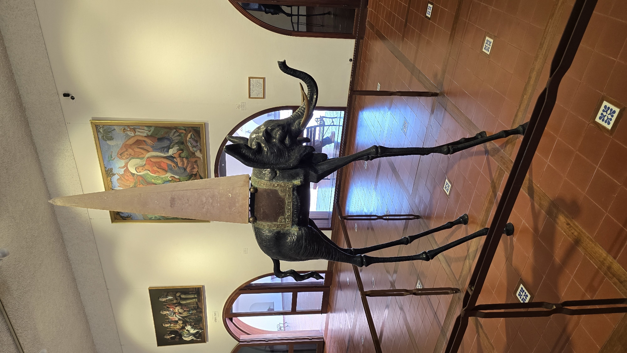 The image shows a surreal sculpture of an elephant with extremely long, thin legs, standing on a tiled floor within a museum or art gallery. The elephant is dark-colored, likely bronze, and is adorned with a decorative saddle and a tall, pointed, pale pink obelisk-like structure on its back. The sculpture is the central focus of the image. In the background, there are large arched doorways leading to other areas, and paintings hang on the walls. One painting on the left depicts a group of formally dressed people with a dog, and another on the right shows a religious scene with figures in a landscape. The floor is tiled with reddish-brown square tiles interspersed with wooden planks and decorative blue and white tiles. Metal barriers are placed in front of the sculpture, suggesting it is an exhibit. The lighting suggests an indoor setting with overhead lights and natural light filtering in from the doorways. This sculpture is a famous artwork by Salvador Dalí and is located at the Dalí Theatre-Museum in Figueres, Spain, not in Hof HaCarmel Regional Council, Israel.