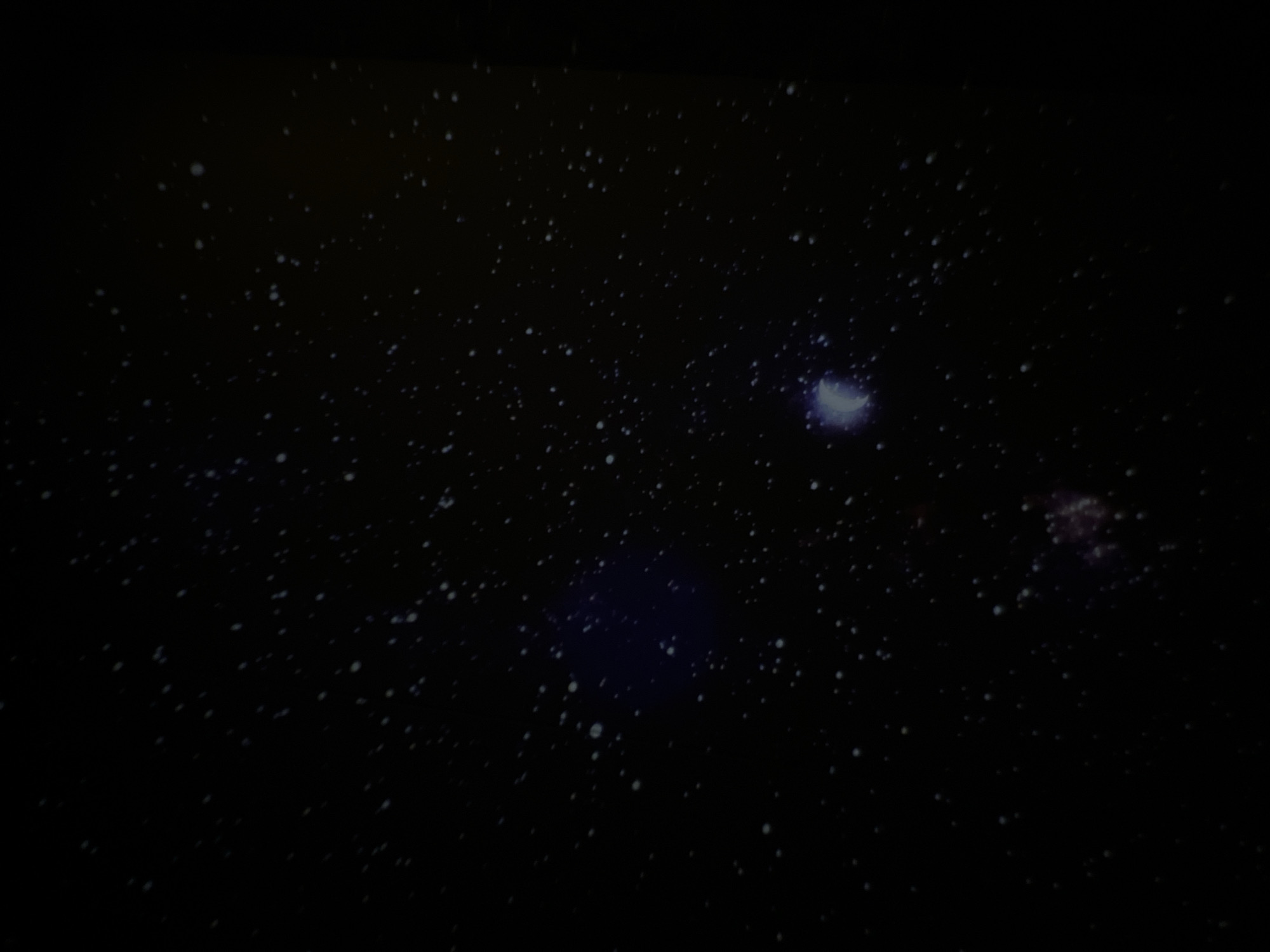 The image depicts a dark night sky with numerous stars visible as small, white dots scattered across the black background. There is a larger, nebulous area of light toward the upper right, with another similar but smaller area of light further down, towards the bottom right. A larger, diffused area of bluish light is located towards the bottom-middle of the image. There is no indication of this image being taken in the city of Chaikovsky, Russia.