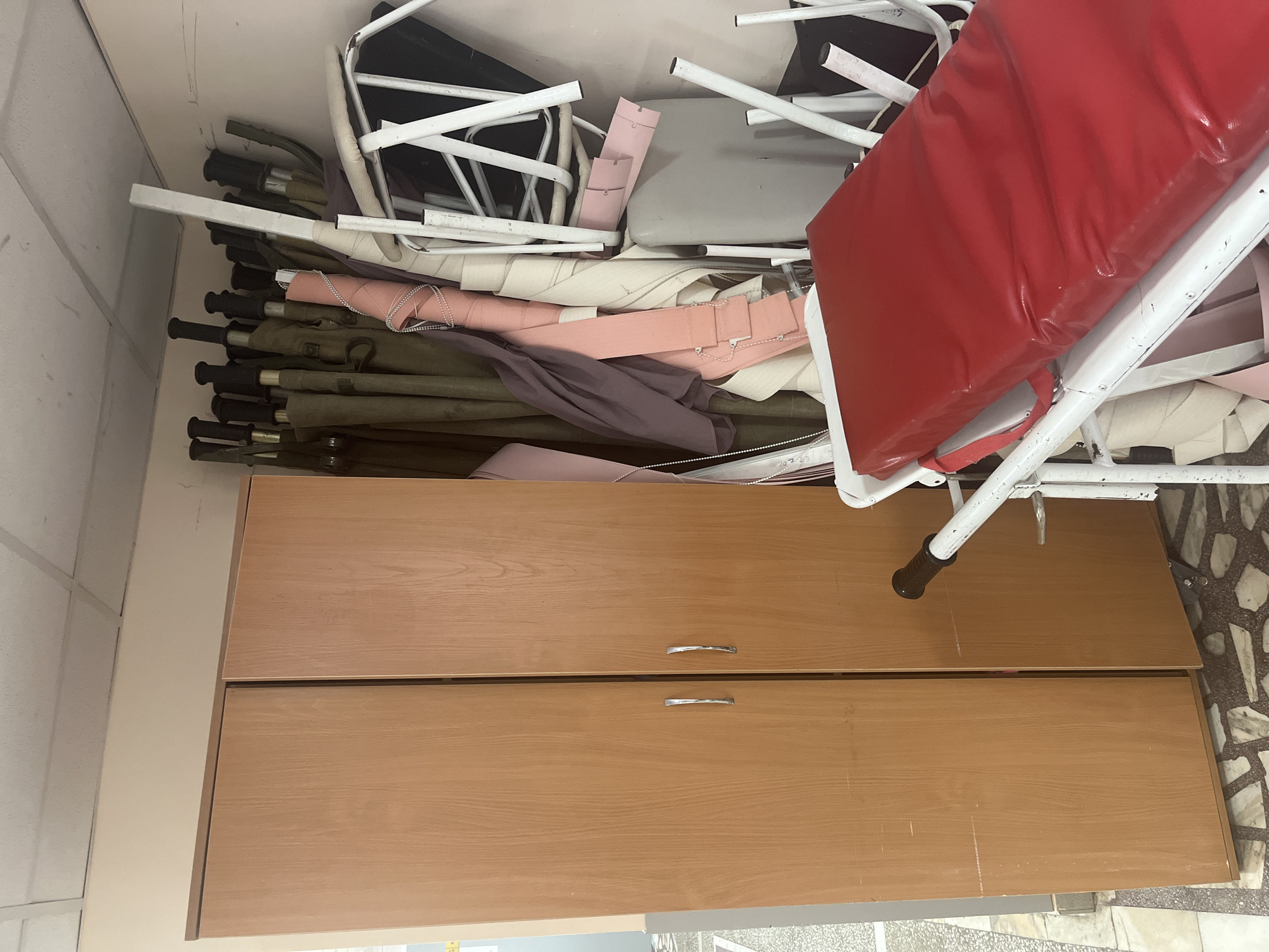 The image depicts a storage room or closet in an indoor location. Dominating the foreground is a tall wooden cabinet with two drawers, each equipped with a silver-colored handle. The color of the cabinet is a light brown, resembling the shade of oak wood. Atop the cabinet is a disorganized collection of various items, including what appears to be stacked folding beds or cots, along with several folding chairs. The furniture and items suggest a storage space within a building, likely intended for holding furniture or supplies. The presence of multiple beds and chairs may indicate that it's a storage area in a public setting such as a school, hospital, or community center. No people are present in the scene. The room is located in the city of Unknown, Unknown.