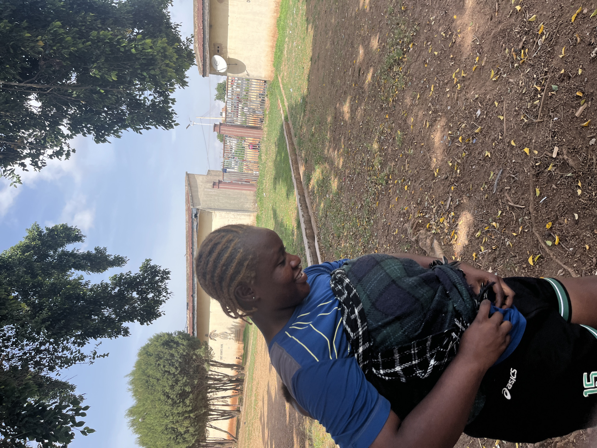A young adult stands in a grassy, sparsely treed area in Gura Top, Nigeria, dressed in a blue shirt with yellow stripes on the sleeves, black shorts with a white "Asics" logo and the number "15," and a plaid blanket draped over their shoulders. The ground is covered in dirt and scattered leaves. In the background, buildings are visible, along with a metal fence and a satellite dish attached to one of the buildings. The sky is light blue with some clouds.