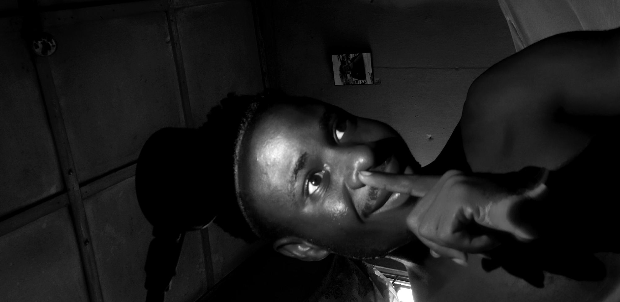 A black and white, medium shot, eye-level photograph shows a young Black male from the chest up, looking to his right and placing his index finger over his nose in a "shushing" gesture. He is wearing a dark, rounded object on his head, possibly a hat or headphones, that casts a shadow over his forehead. His hair is styled with a faded cut around his hairline. He has a light mustache and a playful expression with his eyes wide and a slight smile. His skin is shiny, possibly from sweat or oil, and is well-lit from the front, creating strong highlights and shadows.
The background is dark and appears to be an indoor setting. The ceiling is visible, constructed of dark panels with visible wooden supports. To the right of the subject, on the wall, a small framed picture is hanging. The picture depicts a person sitting and looking towards the camera, though the details are unclear due to the resolution and lighting. The overall lighting suggests a single light source from the front of the subject.
The scene is likely in a home or personal space within Narayi, Nigeria, given the context provided. The image is a self-portrait, suggesting the subject is taking the photo himself, indicated by the angle and framing.