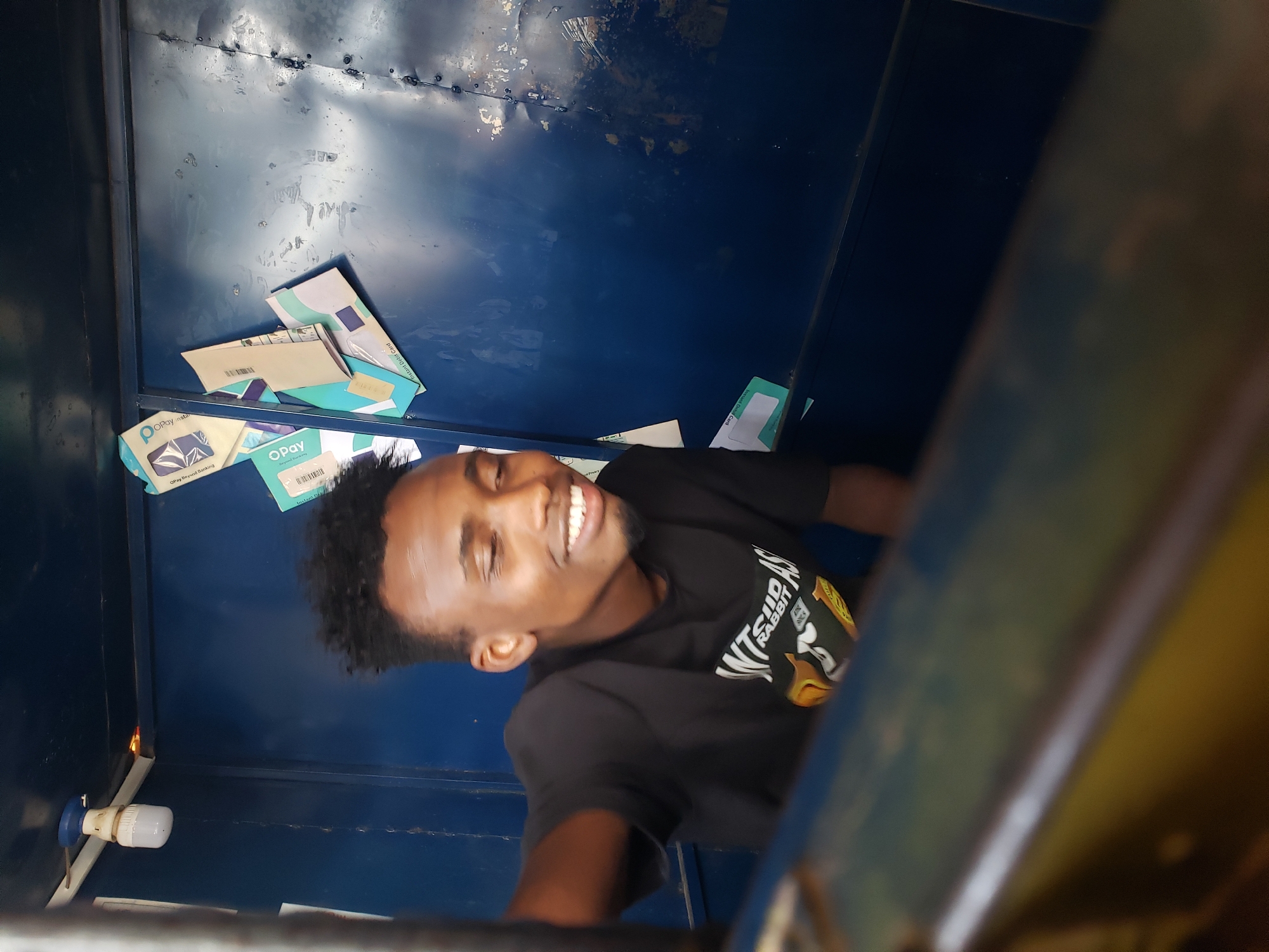 A young Black man with dark, curly hair is smiling and looking to his right, his eyes closed. He is wearing a black t-shirt with white text and an image of a cartoon character. The man is inside a small, blue-painted enclosure, possibly a booth or kiosk, with a light fixture attached to the ceiling. Scattered on the back wall of the enclosure are several paper envelopes and cards, some with the "OPay" logo visible. The enclosure appears to be weathered, with some rust and discoloration on the walls. The foreground is blurred and shows the edge of a yellow and blue structure. The scene is likely in Jos, Nigeria, suggested by the OPay branding, a mobile payment service popular in the region.