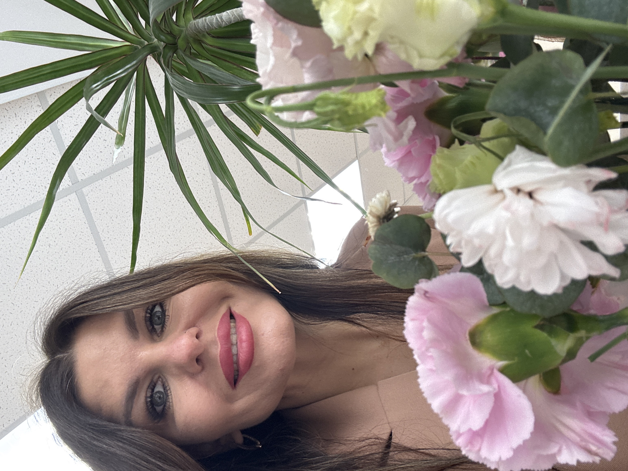 The image depicts a woman with long brown hair, grey eyes, and pink lipstick. She is smiling and looking at the camera from below, with the image tilted. A plant with long green leaves is visible at the top left. Pink, white, and yellow flowers are in the foreground, partially obscuring the woman's face. The background appears to be a white tiled ceiling. The location and action are not provided in the image.