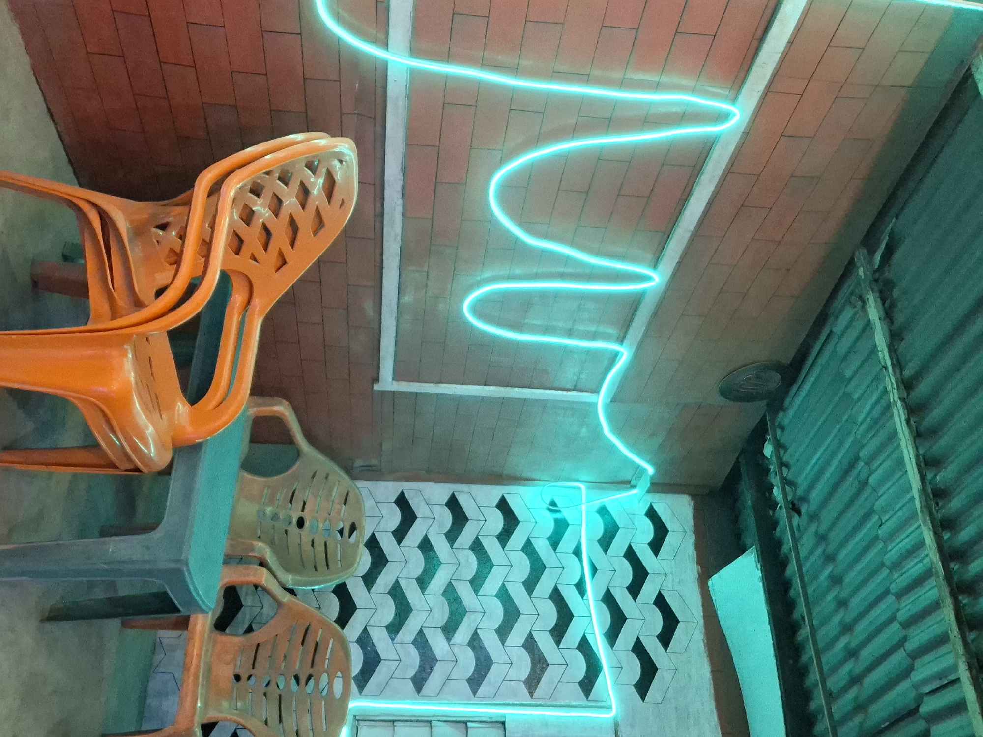 In Keudegeudong, Indonesia, an outdoor seating area is depicted. Three plastic chairs, two orange and one tan, are arranged around a small, teal-colored table. The chairs are stacked in front of a brick wall. Above the brick wall, a bright cyan neon light fixture is installed, forming a wave-like pattern across the wall and continuing onto a patterned wall with geometric shapes. The ceiling is made of corrugated metal, illuminated by the cyan light. The ground is a concrete floor. The scene appears to be in a public space, possibly a cafe or restaurant. There are no people visible in the image.