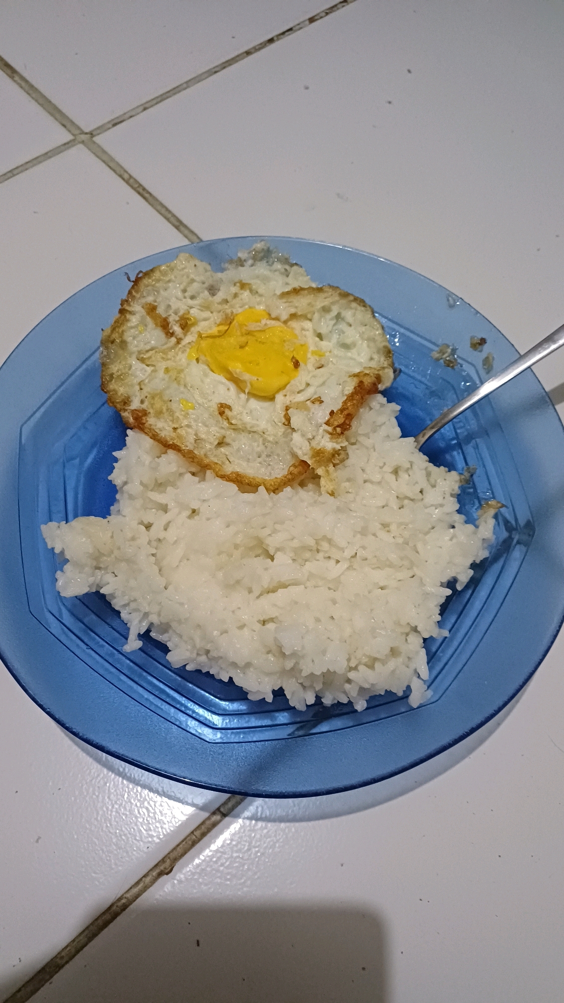 A close-up, overhead shot shows a blue octagonal plate of food placed on a white tiled surface. The plate contains a mound of fluffy white rice and a fried egg with a fully cooked yolk, golden brown around the edges. A silver fork is visible on the right side of the plate, partially submerged in the rice. The white tiles in the background have dark grout lines forming a grid pattern. The scene is well-lit, highlighting the textures of the food and the reflective surface of the plate. This image was taken in Gunung Putri, Indonesia.