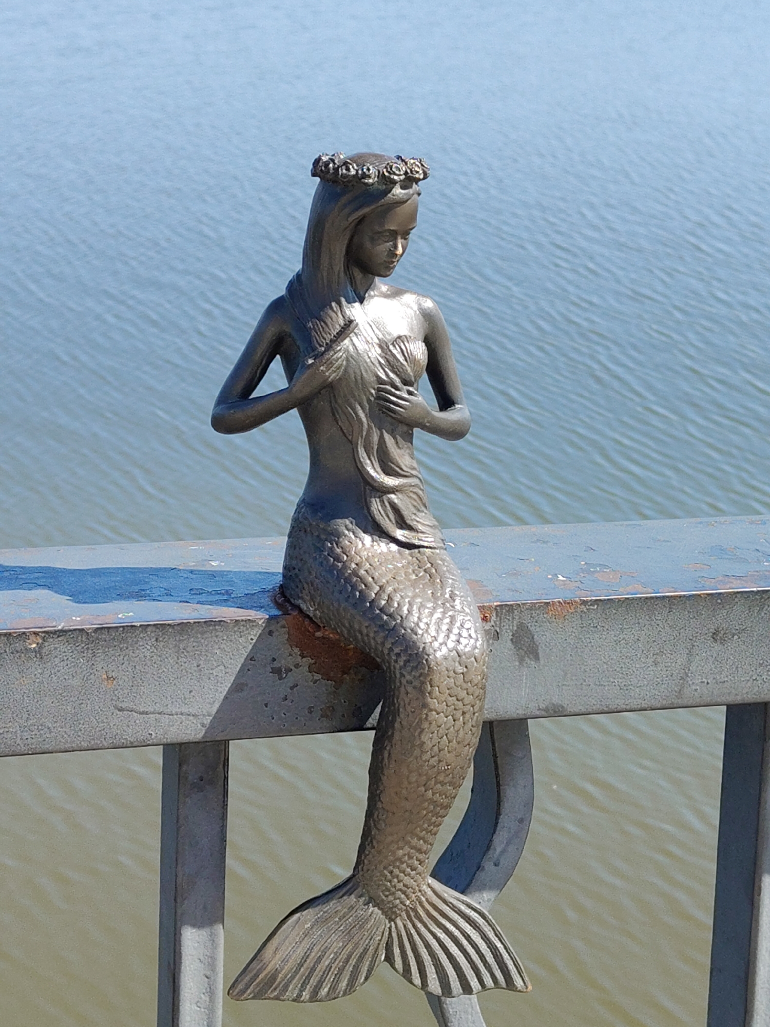 A statue of a mermaid sits on a bridge railing. The statue is made of a dark metal. The mermaid is wearing a floral crown and holding a shell to her chest. The bridge railing is gray. Behind the statue and bridge is a body of water with small waves. The location of the statue is in the city of Unknown, Unknown.