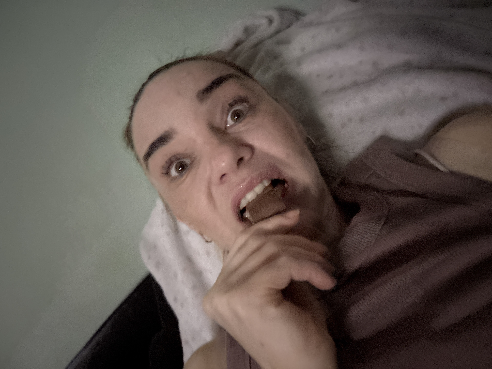 In an indoor setting, a person with wide eyes and an open mouth is seen close-up, biting into a chocolate bar. They are lying on a bed with white patterned bedding and a grey wall in the background. The person is wearing a mauve top. The image is taken from a low angle, with the person's face dominating the frame.